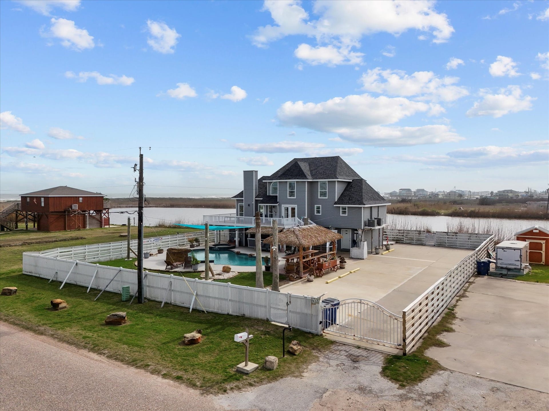 319 Francis Cove, Surfside Beach, TX 77541 This waterfront home has a large fenced yard, outdoor pool, and a patio area with a tiki-style structure. The house has multiple stories and a gray exterior. A detached shed and ample driveway space are also visible, offering plenty of room for parking.