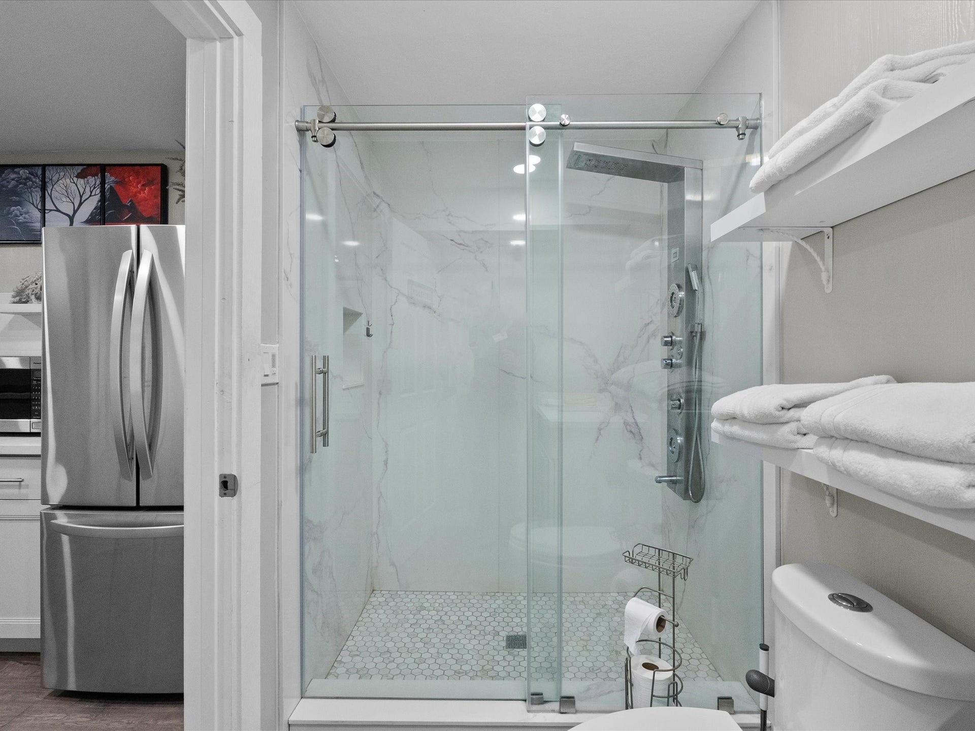 319 Francis Cove, Surfside Beach, TX 77541 This modern bathroom features a sleek glass-enclosed shower with a rain showerhead and marble-style walls. It includes a dual-flush toilet and built-in shelving for towels. Adjacent is a glimpse of a contemporary kitchen with stainless steel appliances.