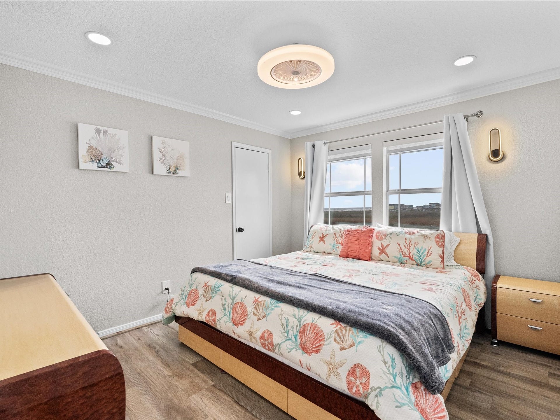 319 Francis Cove, Surfside Beach, TX 77541 This inviting bedroom features a comfortable bed with a coastal-themed bedspread, soft lighting, and natural light from two large windows. The neutral walls and wood flooring create a calming atmosphere, complemented by modern wall sconces and simple artwork.