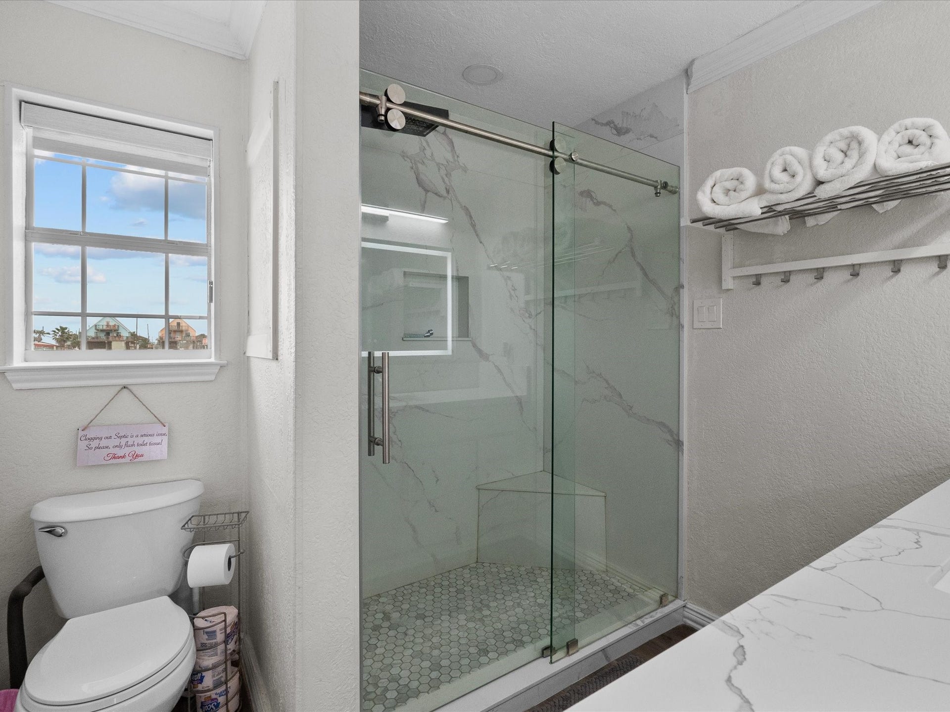 319 Francis Cove, Surfside Beach, TX 77541 This bathroom features a modern design with a glass-enclosed shower, marble-patterned tiles, and a sleek countertop. It has a window for natural light and open shelving for towels.