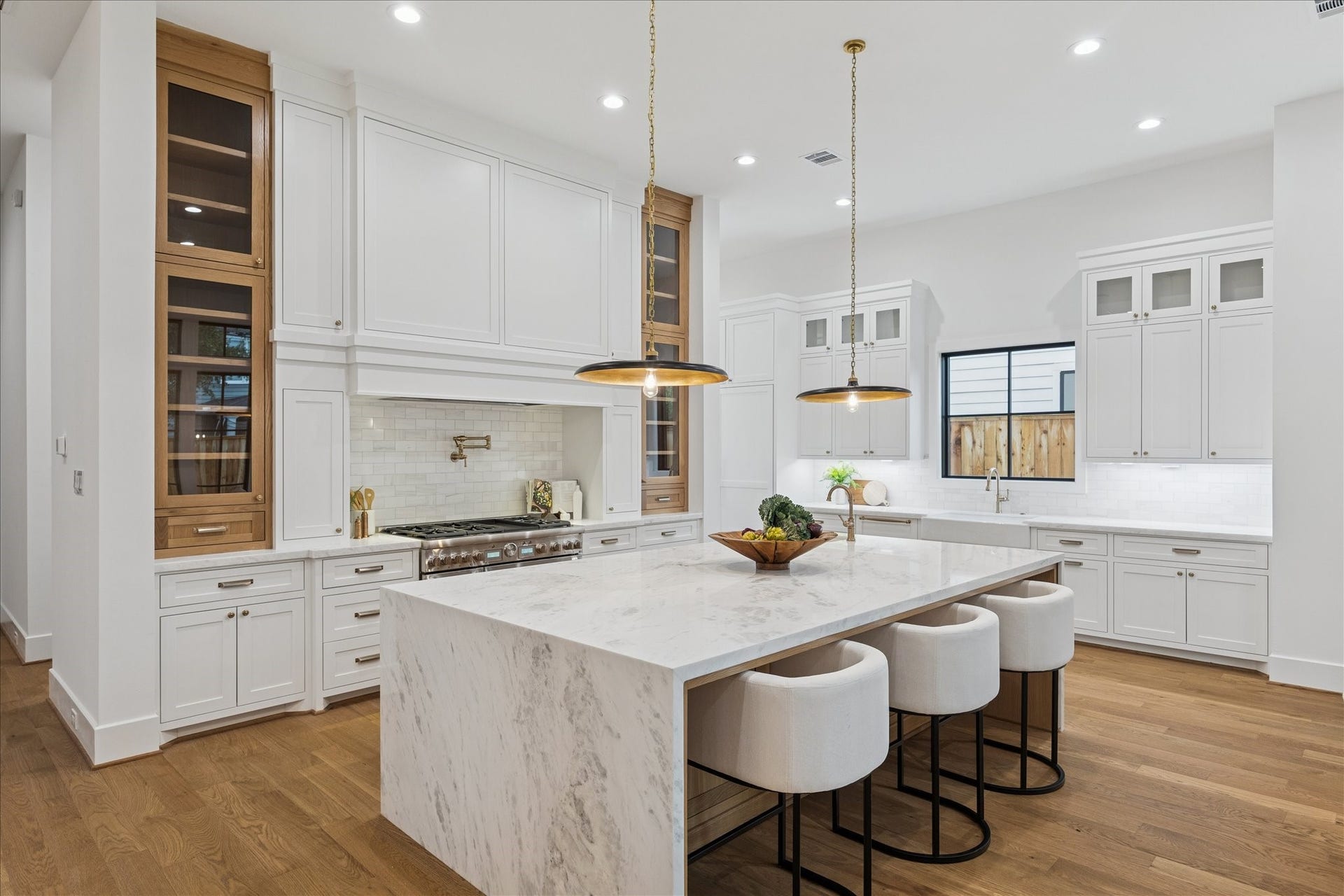 Floor-to-ceiling millwork frames a professional-grade Thermador range beneath a sculpted hood, flanked by vertical glass cabinets for elegant display. Brass pendants cast a warm glow across the marble surfaces, while the Pella window package and open layout ensure a bright, functional cooking space. *Previous Project*