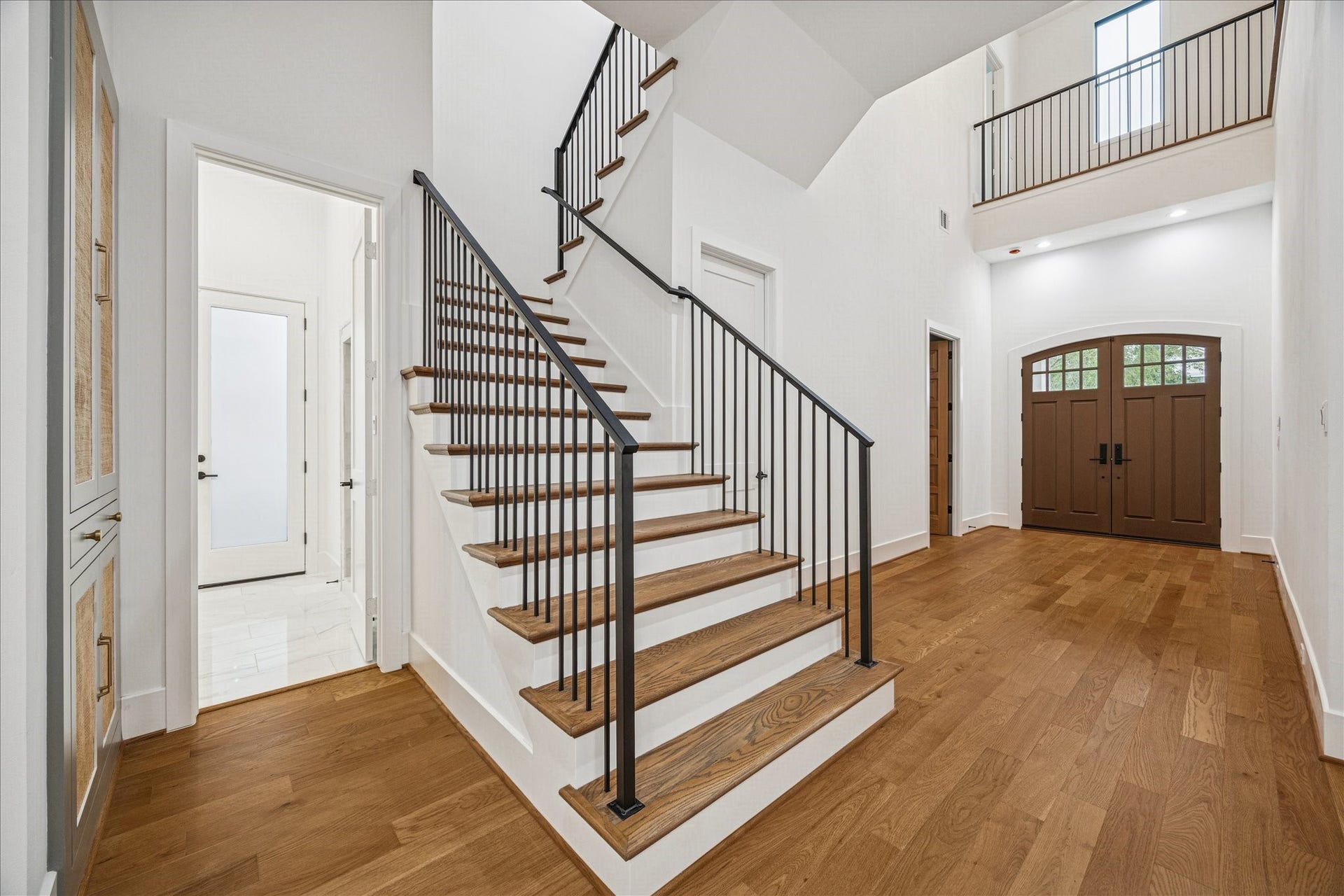 The wide gallery hall establishes elegant flow toward the homes core living spaces. Tall ceilings emphasize openness, while continuous oak flooring adds warmth and cohesion. Iron railing introduces sculptural contrast. Raffia-inset cabinetry provides functional storage and textural depth, enhancing the homes balance of refined design and everyday usability. *Previous Project*