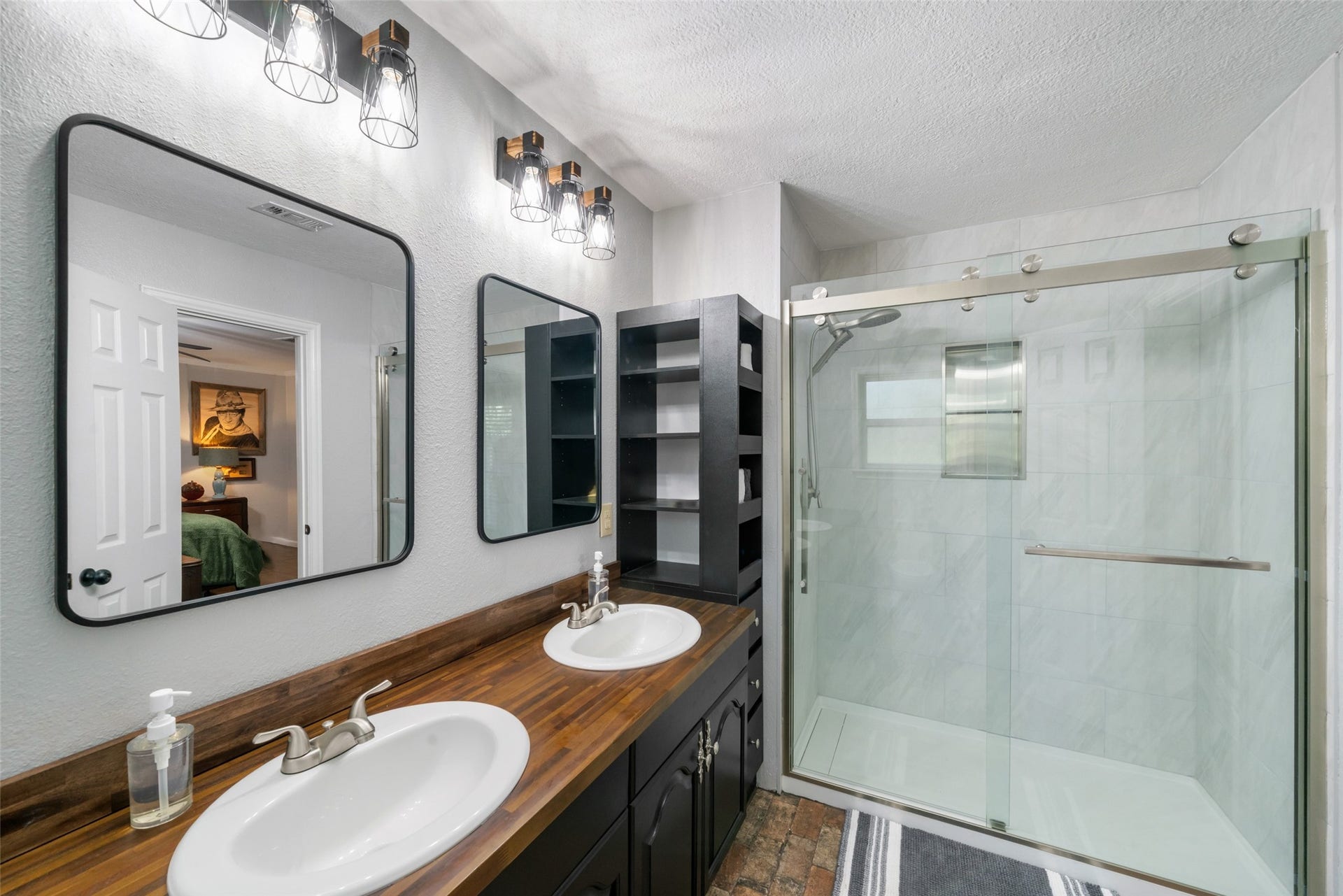 This updated primary bathroom is a true showpiece, spacious, beautifully finished, and designed with both style and function in mind. The large walk-in shower features a sleek linear trough drain, giving it a modern, high-end feel. Decorative lighting adds warmth and character, while the gorgeous double-sink vanity and stylish mirrors create a polished, inviting look. The brick-style tile flooring brings in texture and charm, and the vanity offers plenty of storage, including convenient corner shelving.