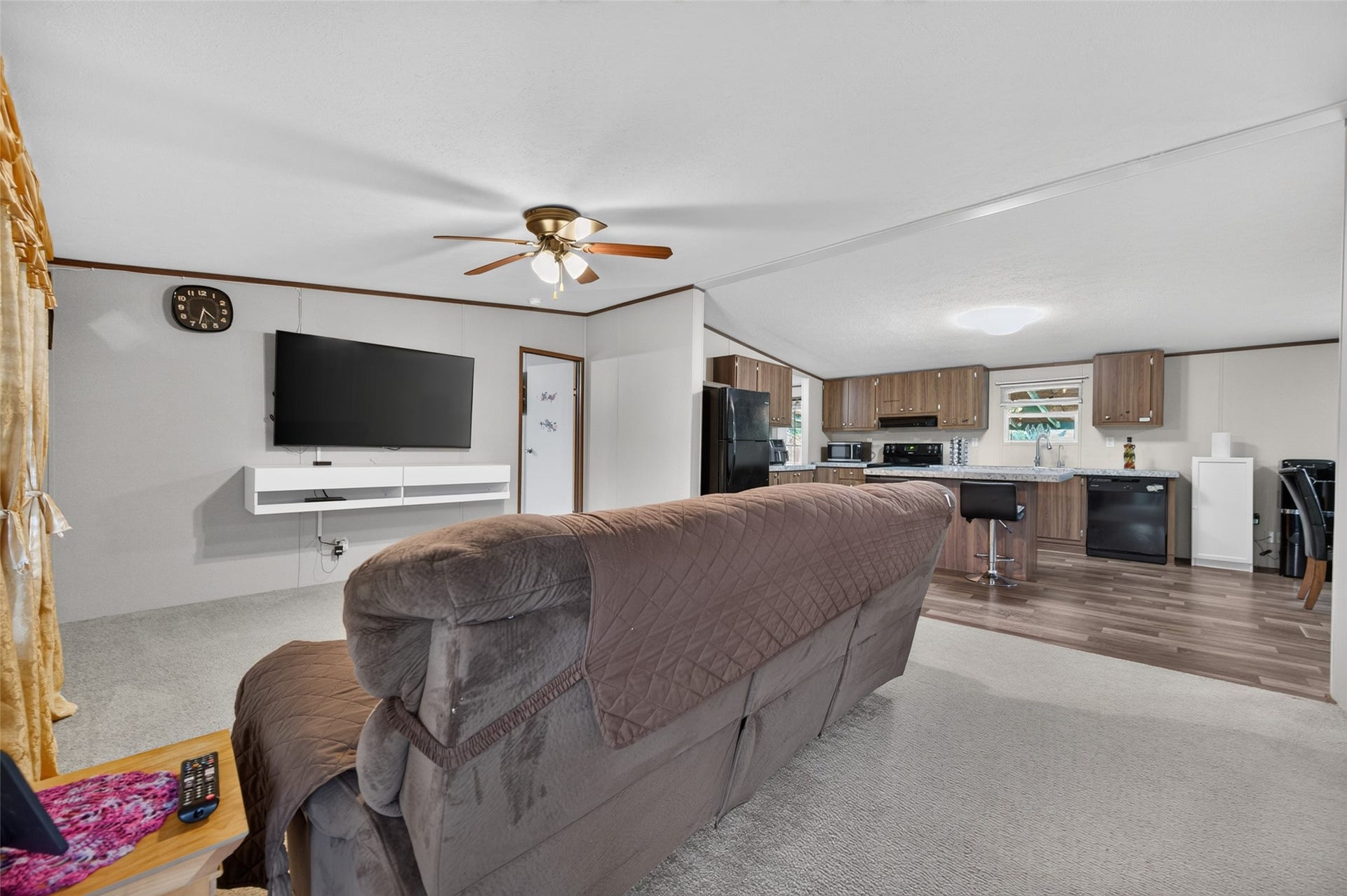 Bright living room with ceiling fan, and mounted TV nook, opening to the kitchen