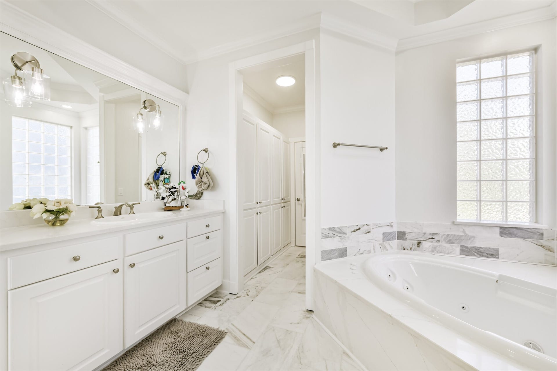 Bathroom featuring a luxurious marble-tiled jacuzzi tub, dual vanities with ample storage, and modern fixtures. Natural light pours in through frosted glass windows, creating a serene and inviting atmosphere.