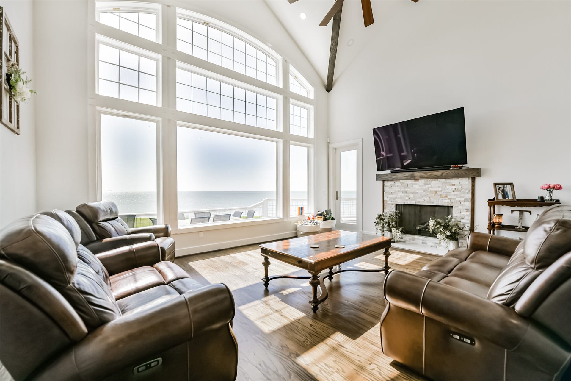 Experience breathtaking ocean views through expansive windows in this spacious living room, featuring a cozy stone fireplace, elegant leather seating, and soaring vaulted ceilings.