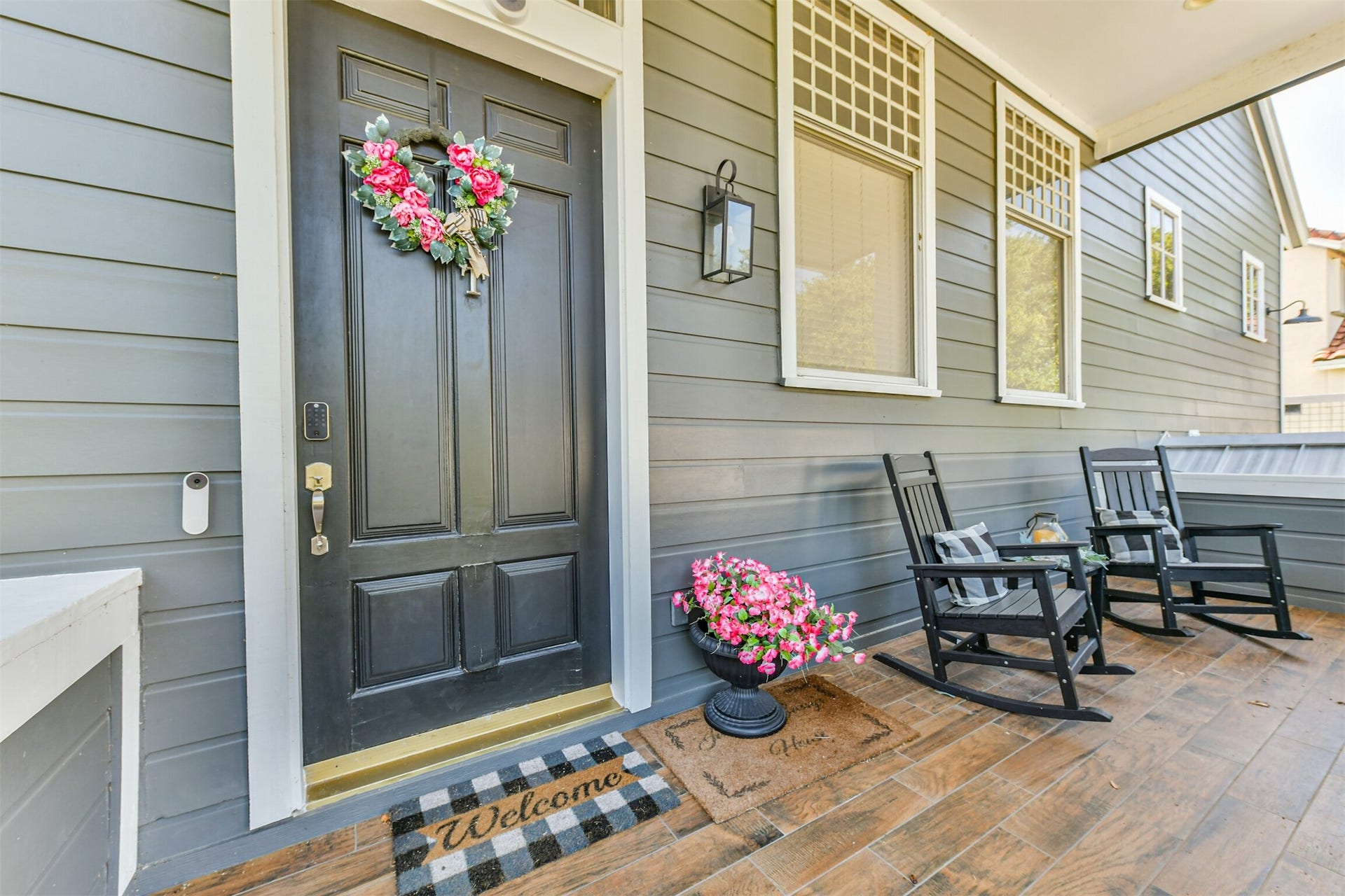 Cozy front porch with elegant dark wood door, adorned with a floral wreath. Relax in the cozy rocking chairs on the inviting wood-tiled floor, surrounded by vibrant flowers.