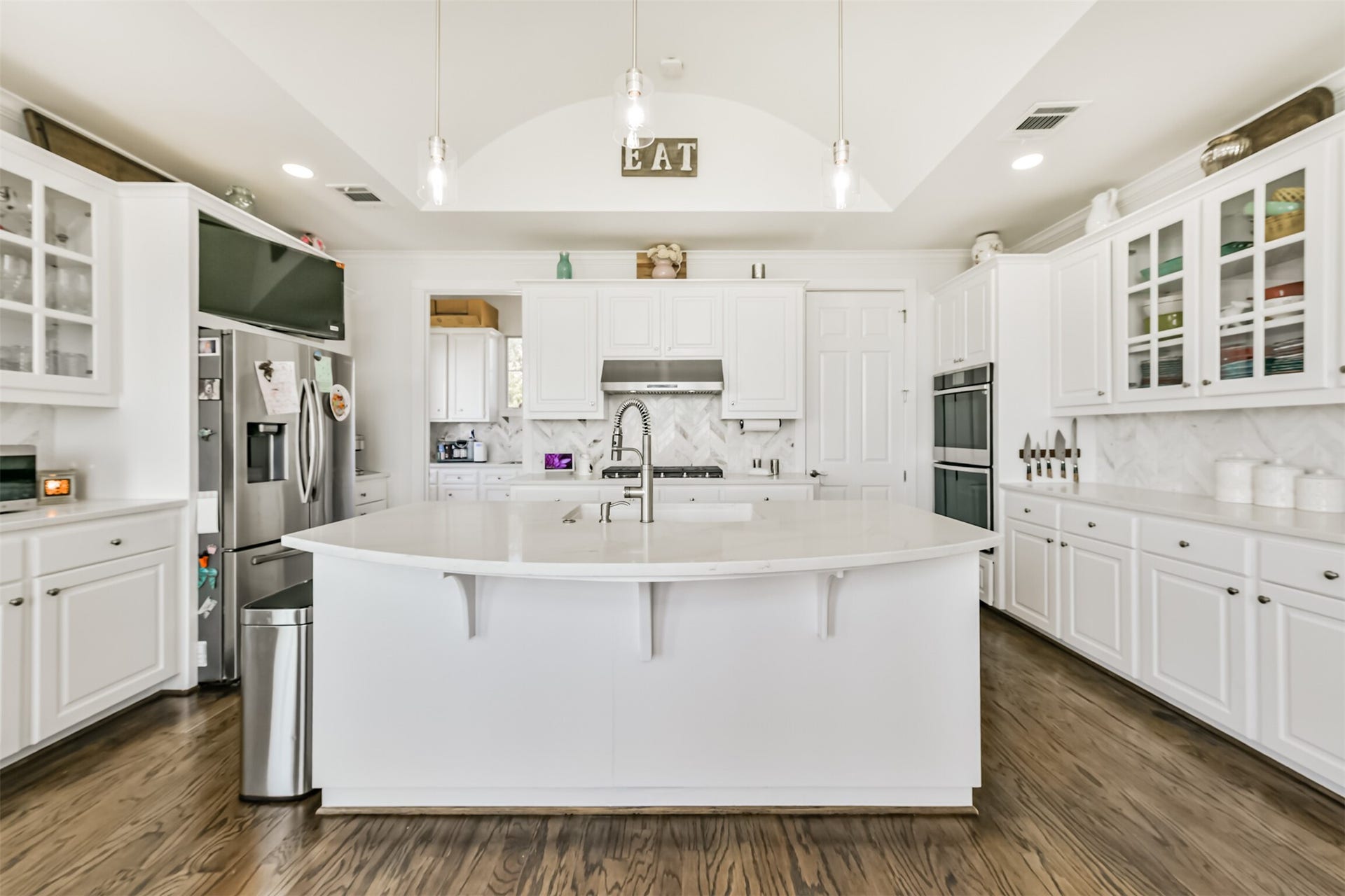 Experience modern elegance in this spacious kitchen featuring gleaming white cabinetry, a large central island with a sink, and stainless steel appliances. Abundant natural light and sleek pendant lighting accentuate the open layout, perfect for culinary creativity and entertaining.