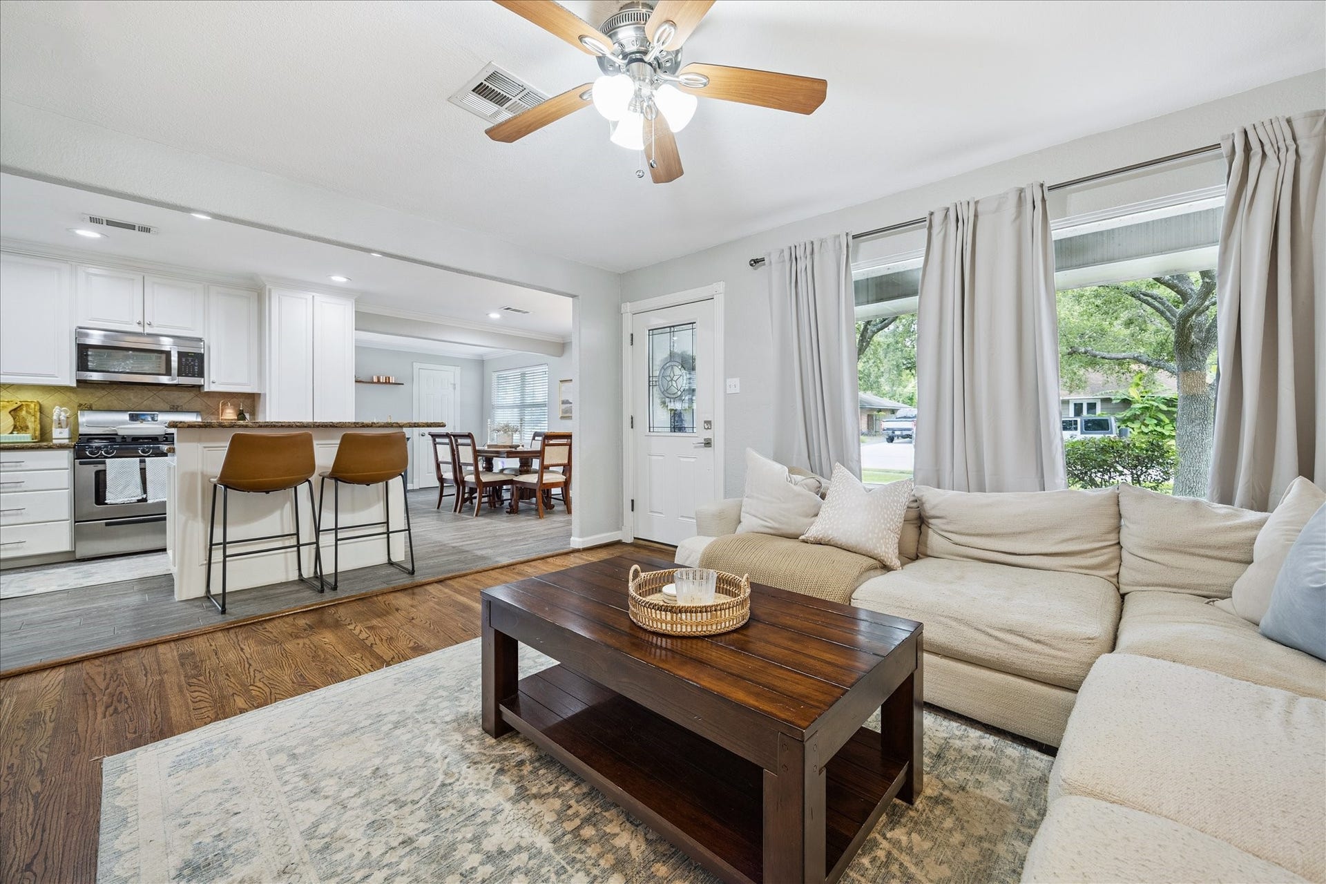 The living room opens seamlessly to the kitchen and dining area, with triple windows bringing in soft natural light and a wide view of the front yard.