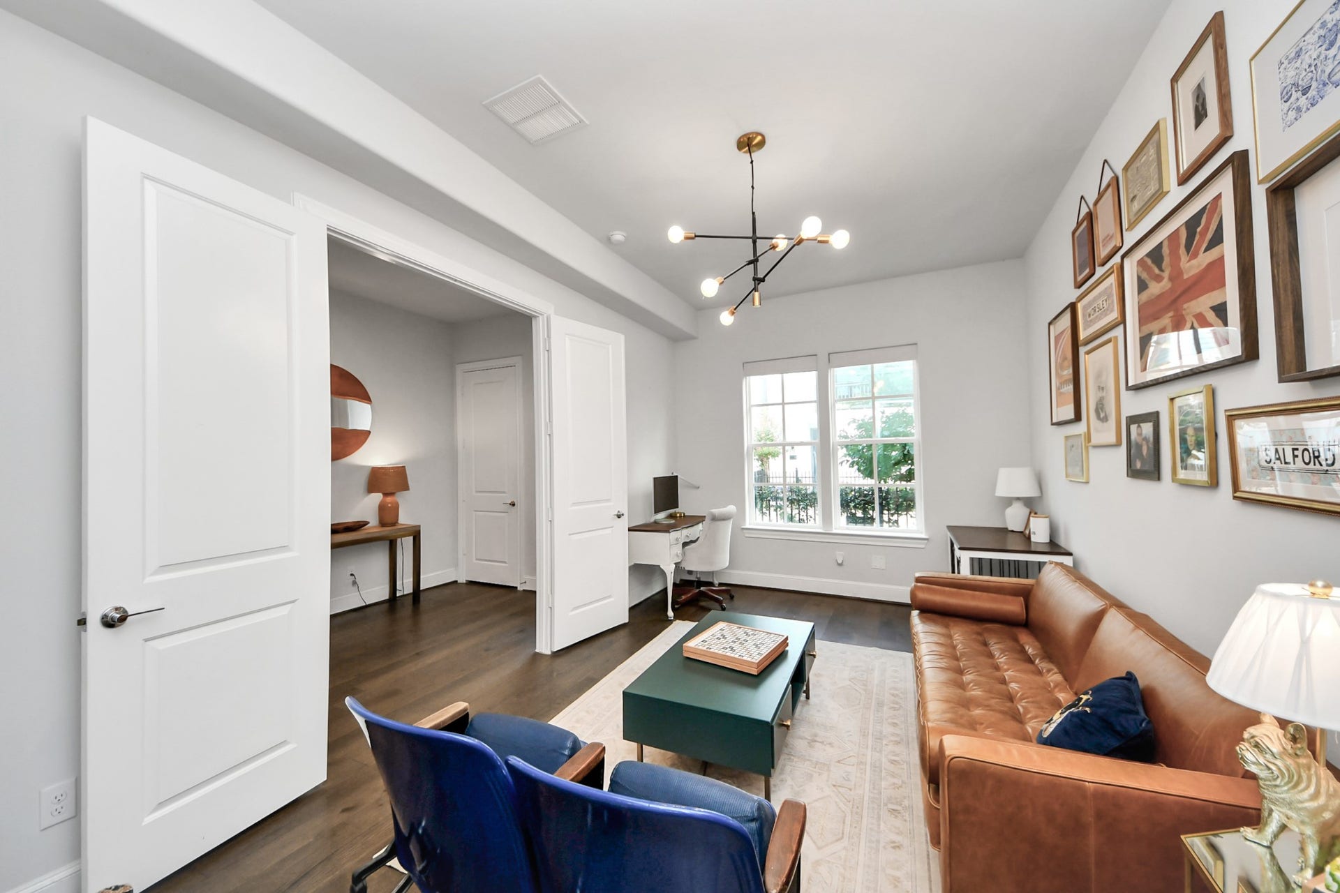 Stylish and inviting living area with a modern chandelier, cozy leather sofa, and a gallery wall of art. The space is complemented by a home office nook with natural light streaming through large windows.