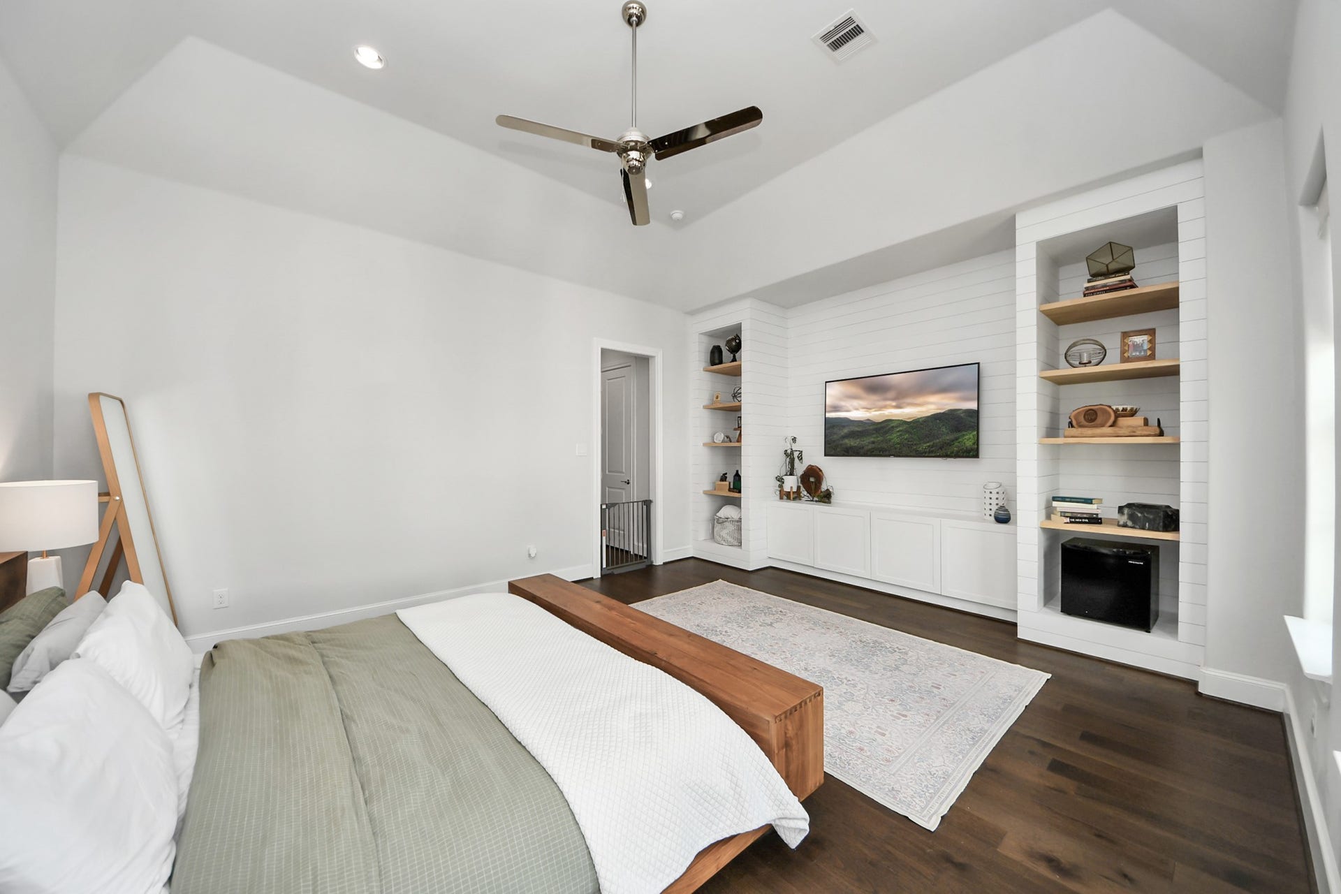 Spacious bedroom with high ceilings, featuring a sleek ceiling fan, built-in shelving, and a mounted TV for a modern, relaxing retreat.