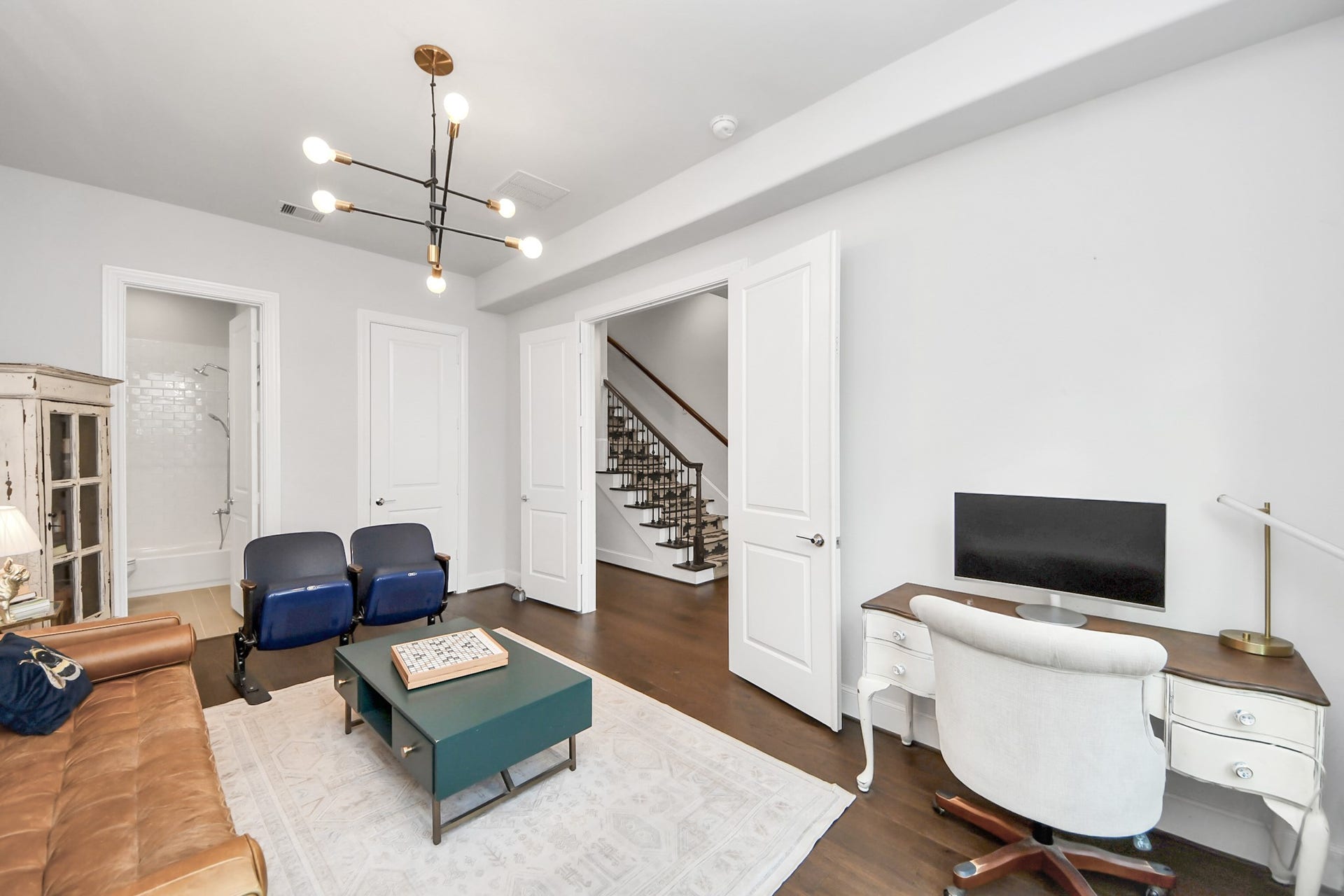 Elegant and versatile room with modern lighting, featuring a cozy sitting area, a functional workspace with a white desk, and easy access to the staircase.