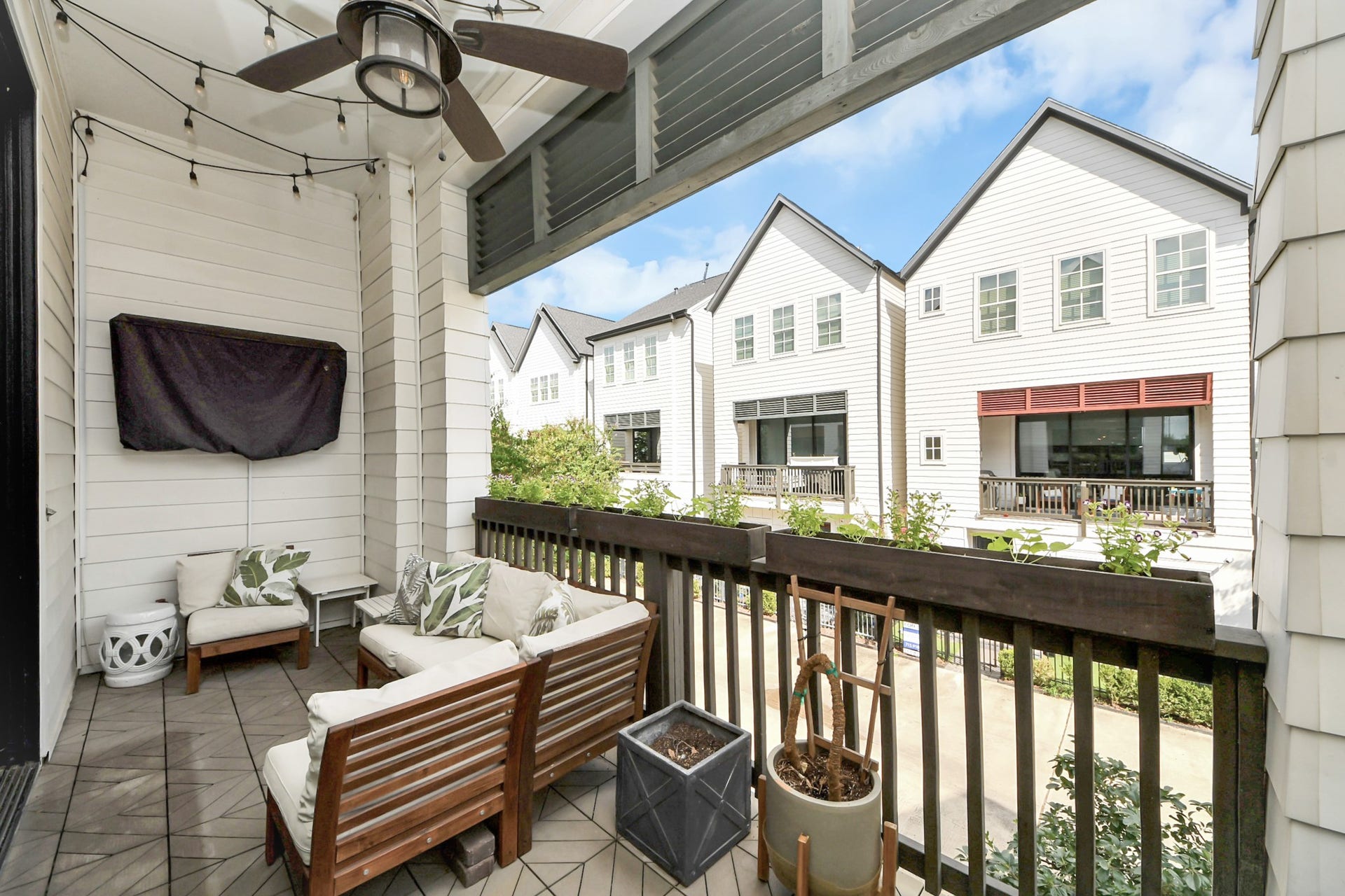 Charming covered balcony with comfortable seating and a ceiling fan, overlooking modern townhomes and lush greenery.