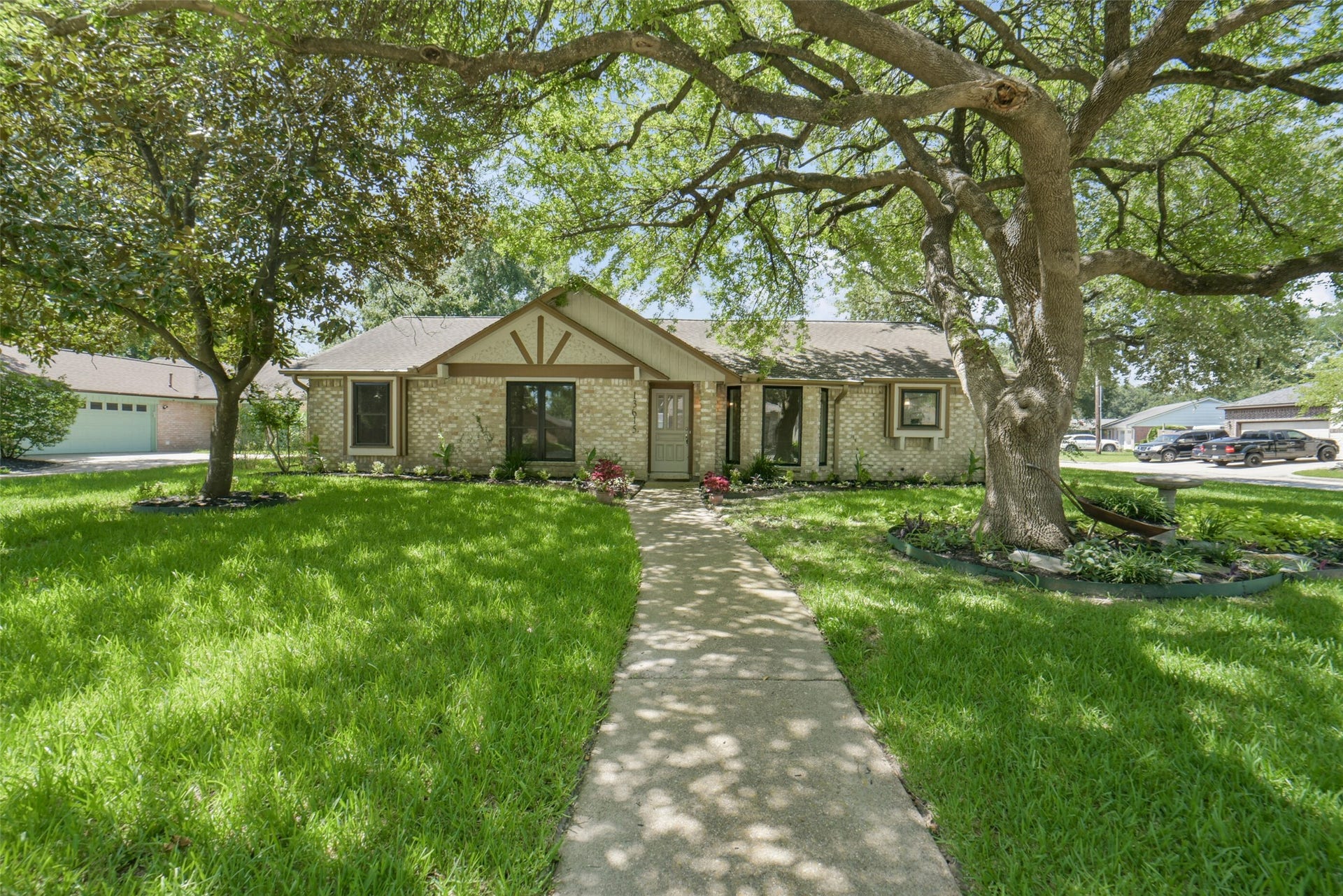 Charming single-story brick ranch home shaded by mature oak trees, featuring a manicured lawn, a long concrete walkway leading to the front entrance, well-maintained landscaping beds, and a vaulted front gable with wood accents  offering classic curb appeal in a quiet residential setting.