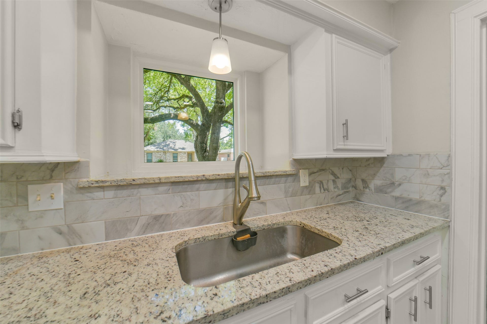 Kitchen sink area featuring a garden window with a picturesque tree view, a stainless steel single-basin sink with gooseneck faucet, granite countertops, marble-look tile backsplash, and a pendant light overhead