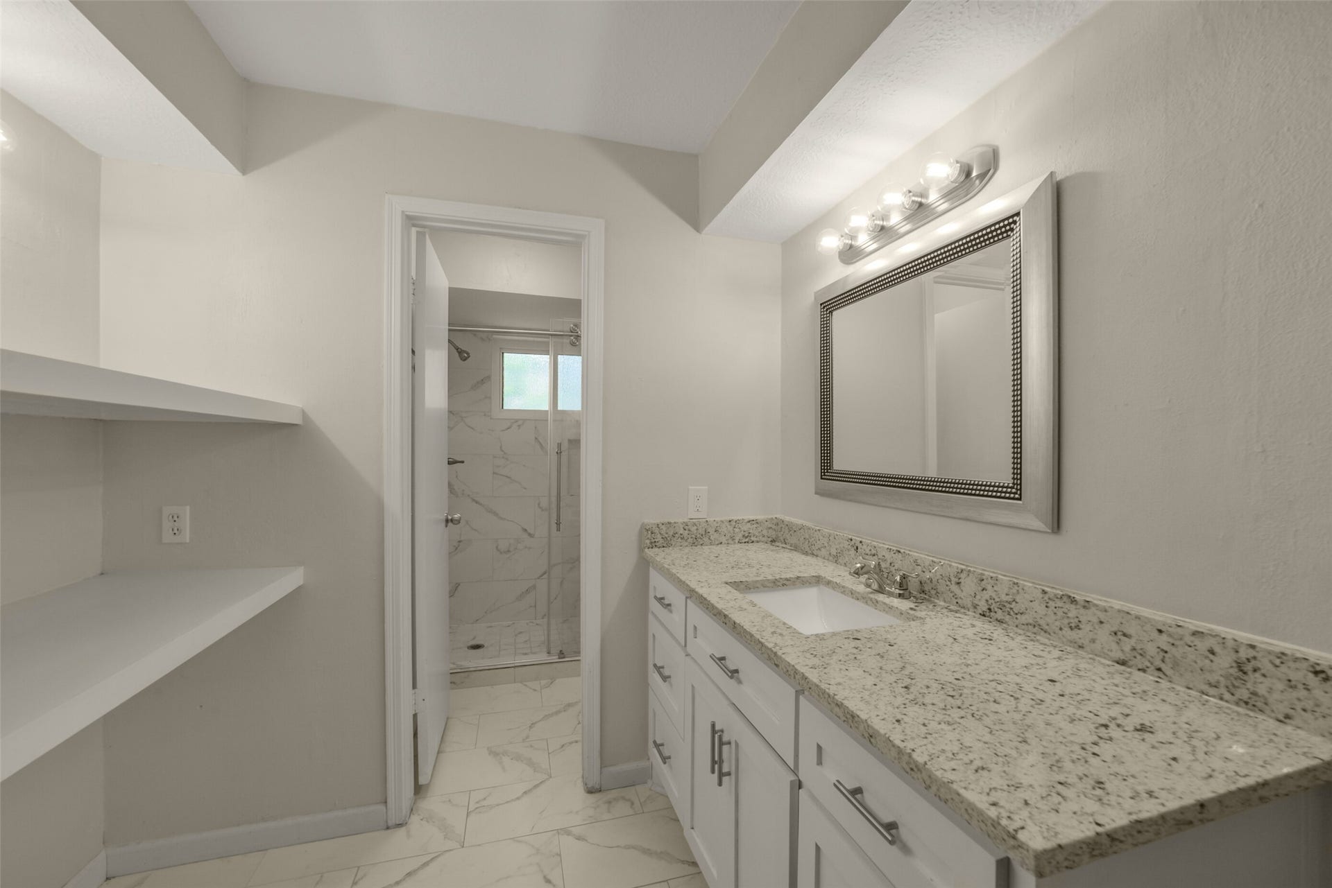 Elegant primary bathroom featuring a granite-topped vanity with white cabinetry, a stylish framed mirror with sleek vanity lighting, and marble-look tile flooring throughout. A separate walk-in shower with glass enclosure and matching marble-look tile surround completes this spa-like retreat.