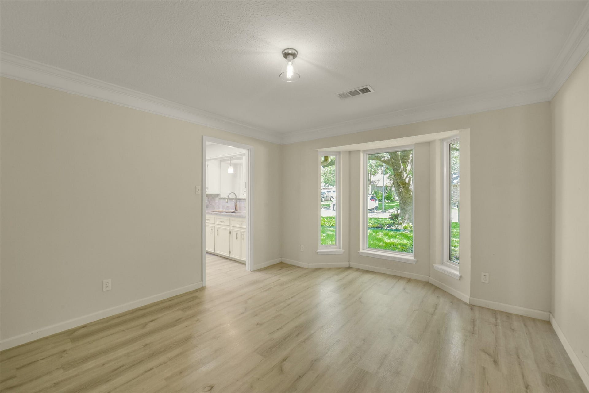 Bright formal dining area featuring an elegant bay window overlooking the front yard, wood-look flooring, crown molding, and a convenient pass-through view into the kitchen  an inviting space for hosting family meals and gatherings.