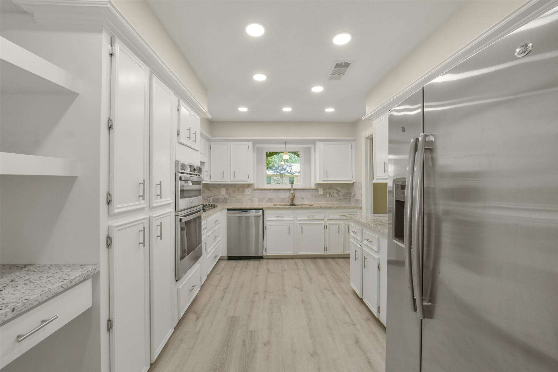 Wide view of the fully renovated kitchen showcasing crisp white cabinetry, granite countertops, a stainless steel refrigerator with ice/water dispenser, double wall oven, dishwasher, and gas cooktop. Recessed lighting, a pendant fixture over the sink, and a garden window bring in natural light, while a spacious pantry with granite counter adds extra storage  a bright, move-in-ready kitchen designed for everyday living.