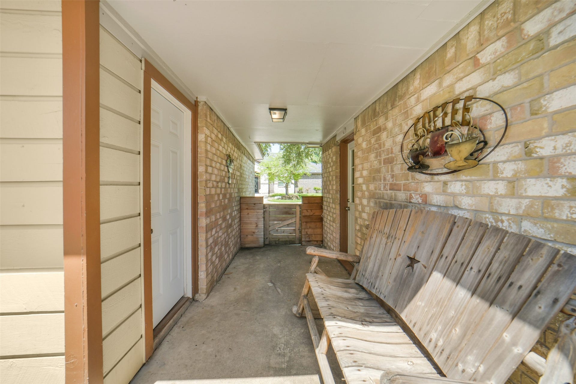 Covered breezeway/porch connecting the home to the garage, featuring a charming rustic bench, brick and wood accents, and a gated side entry  a cozy outdoor spot for relaxing or transitioning between spaces.