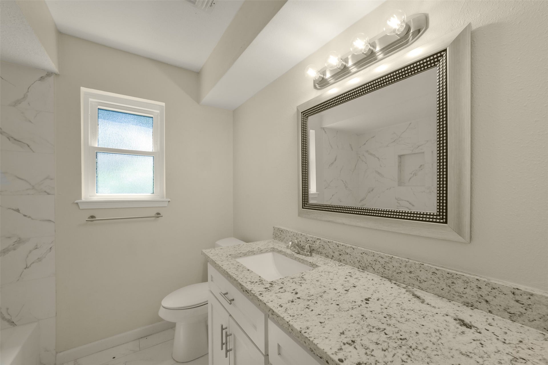 Renovated bathroom featuring a granite-topped vanity with white cabinetry, an updated framed mirror with modern vanity lighting, and marble-look tile surrounding the tub/shower  a fresh, elegant space with a privacy window for natural light.