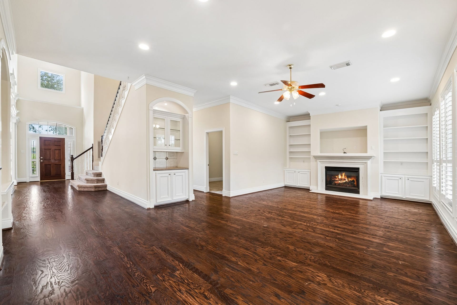 Viewed from breakfast area, showing Texas sized Family room area featuring large windows with elegant plantation shutters, neutral walls, built-in shelves, niche with a focus light and long stretch of wood floor to the foyer area.