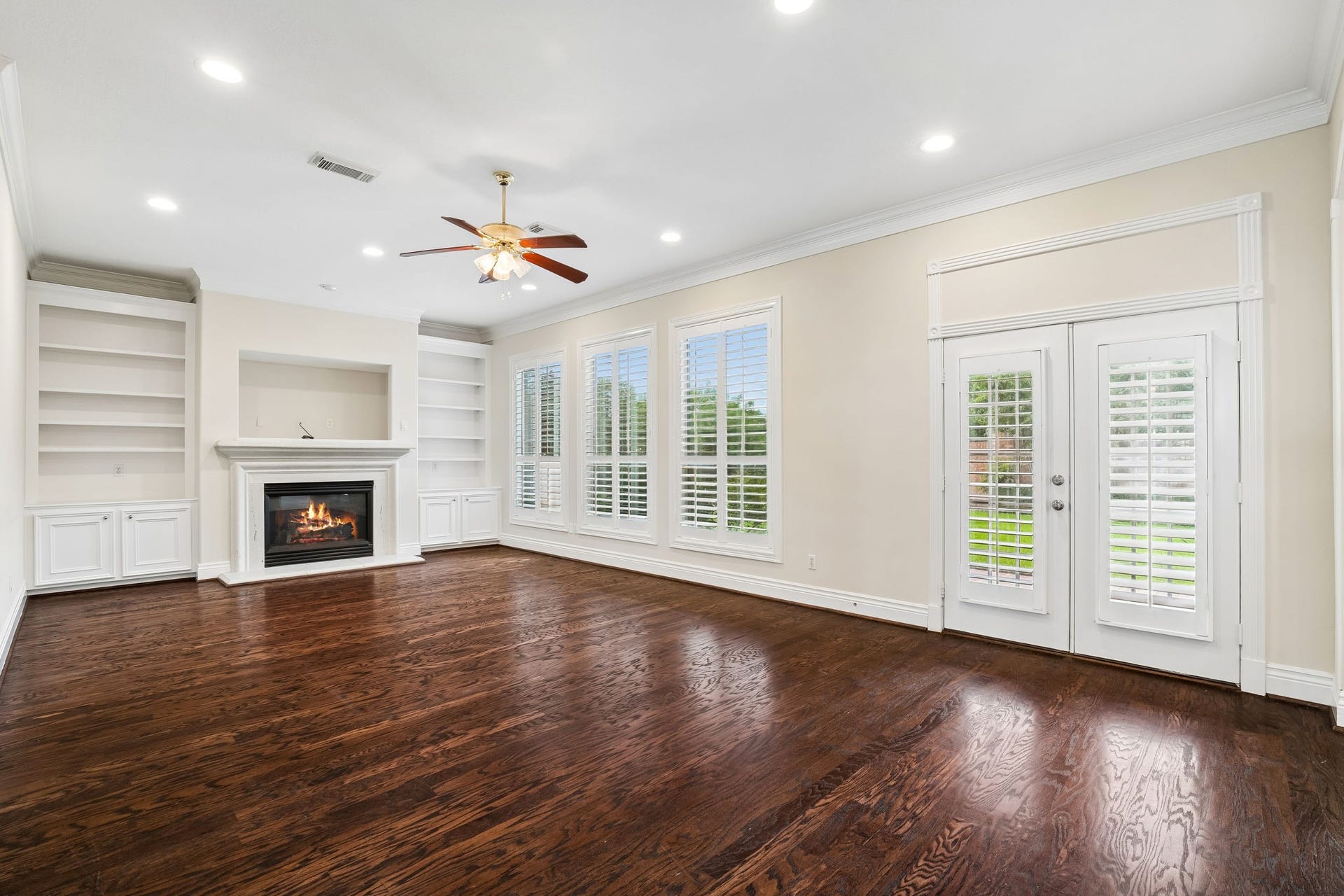 Spacious family room featuring gleaming hardwood floors, a cozy fireplace, and abundant natural light from large windows with elegant shutters. Built-in shelves add charm and functionality.