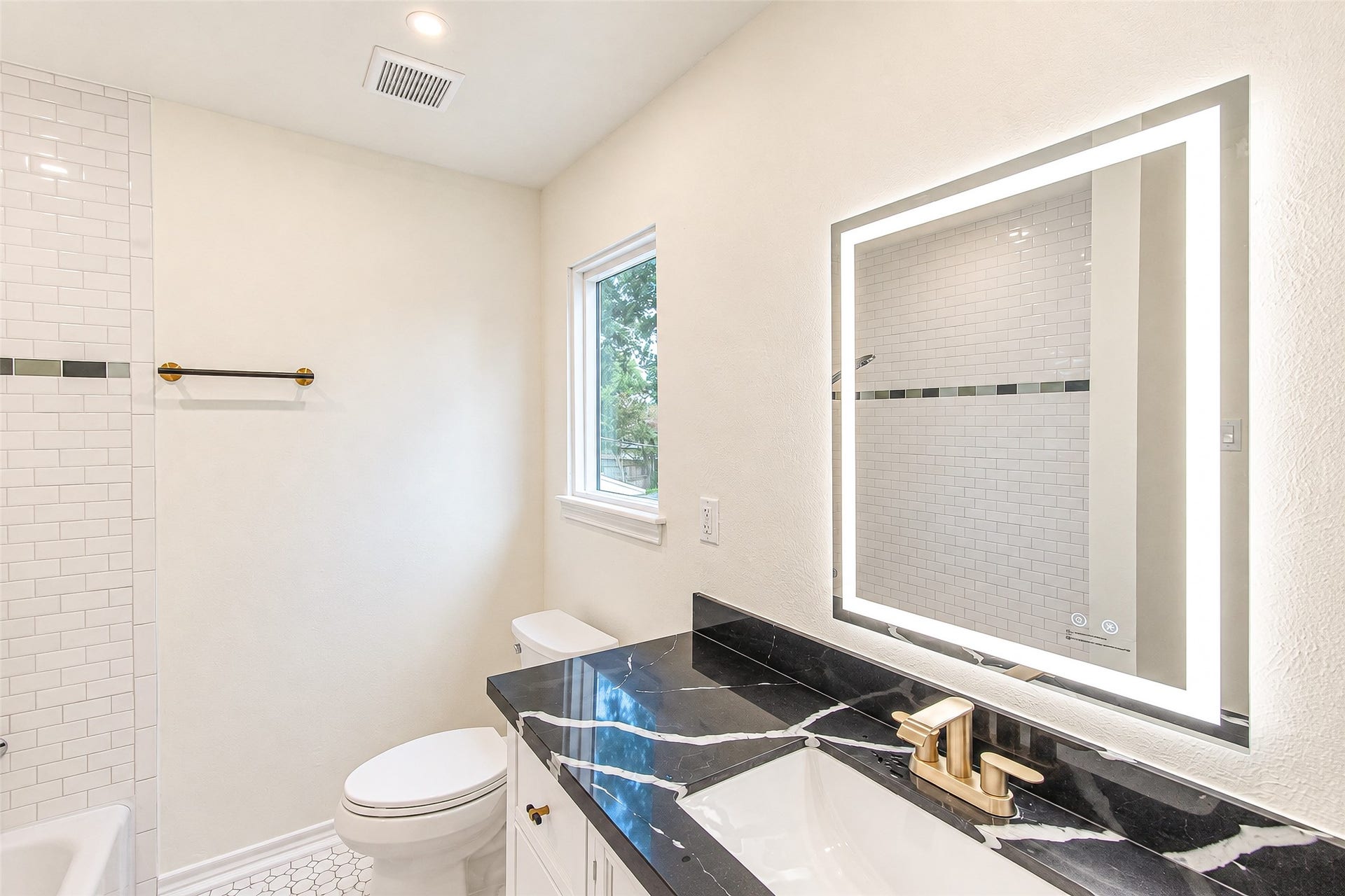 10610 Overbrook Lane, Houston, TX 77042 Luxuriate in the primary bath featuring a sleek black quartz countertop, modern gold fixtures, and an elegant backlit mirror, all complemented by pristine white tiles.