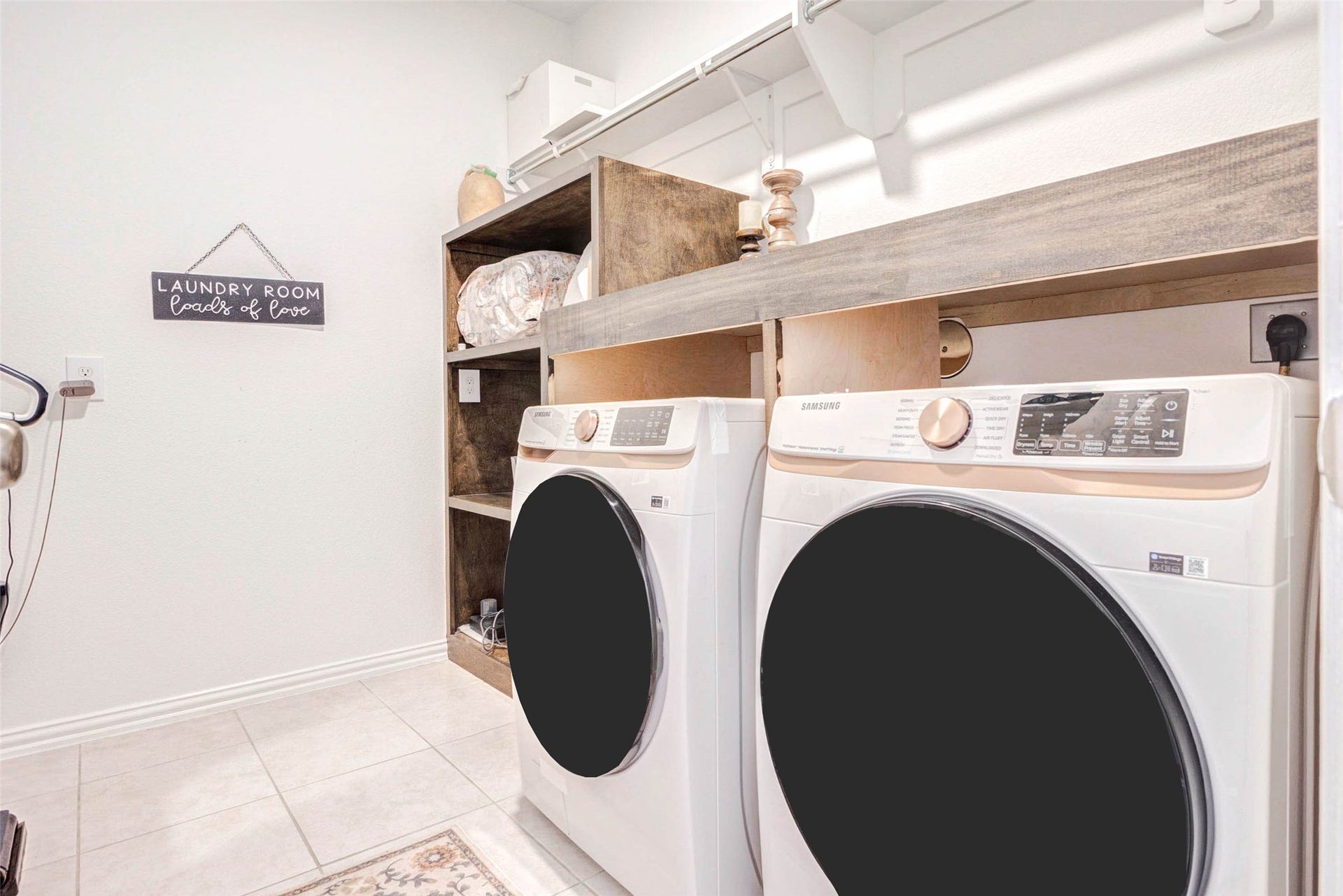 This dedicated laundry room is designed for efficiency and convenience, featuring side-by-side washer and dryer units with overhead shelving for storage. Built-in cabinetry and open shelving provide plenty of space for organization, while the clean layout keeps everything within easy reach. Bright lighting and durable flooring make this a practical and functional space for everyday use.