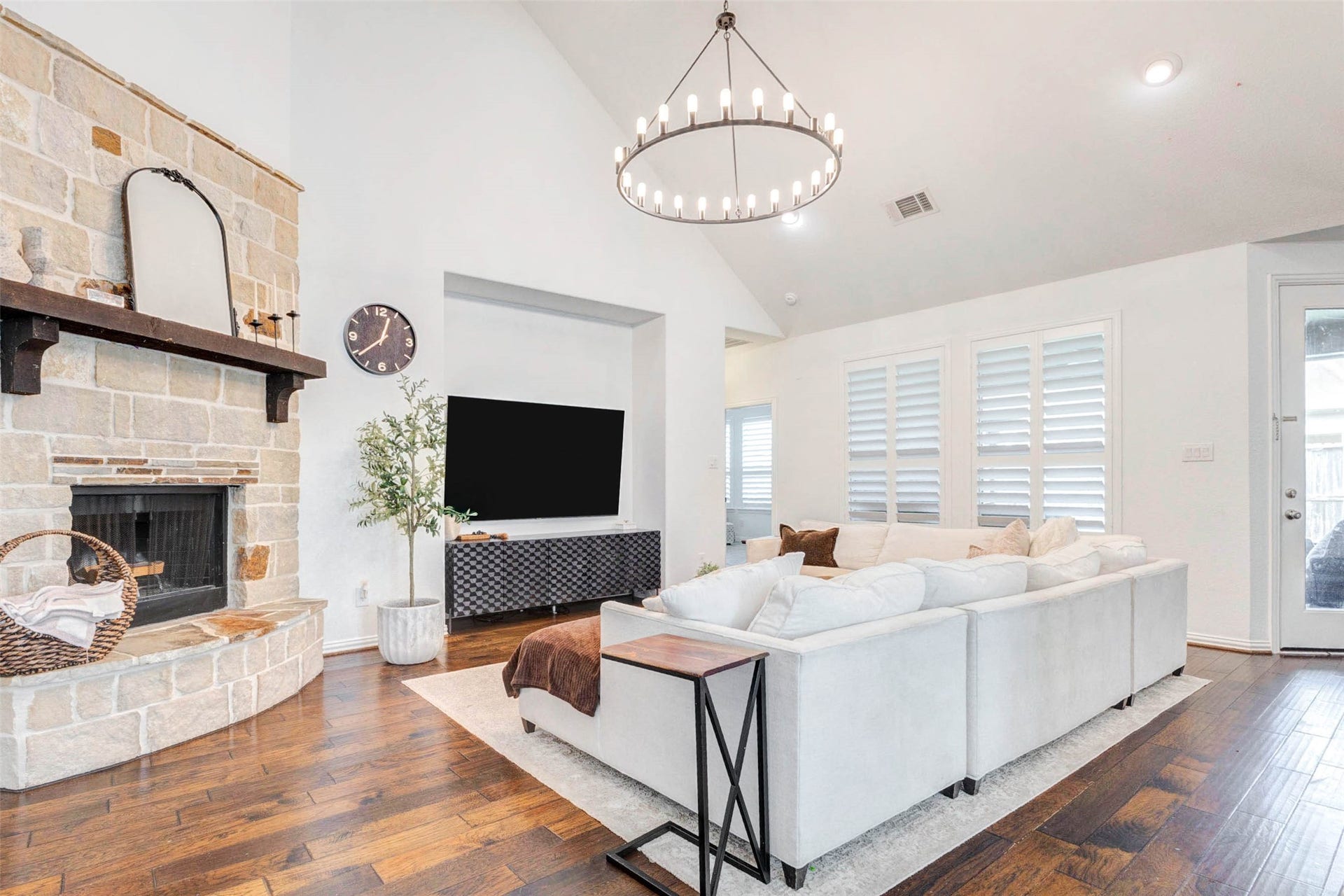This expansive living room is filled with natural light and designed for both comfort and style. Soaring ceilings and a statement chandelier create an airy, open feel, while the stone fireplace adds warmth and character. Large windows with plantation shutters enhance privacy and elegance. The seamless flow into adjacent spaces makes this area perfect for entertaining or relaxing in a beautifully curated environment.