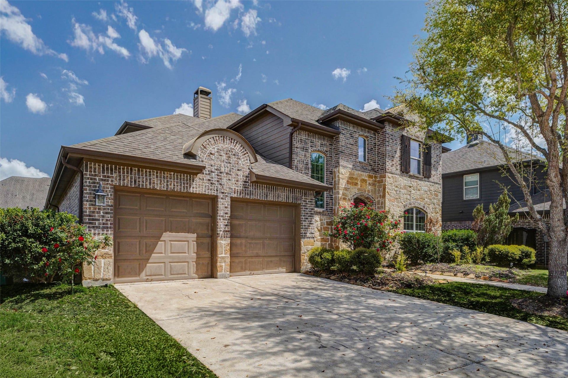 This striking front facade blends functionality with style, featuring a three-car tandem garage and extended driveway. The brick exterior adds depth and character, while well-maintained greenery and flowering shrubs soften the look. The home's architectural details, including arched windows and rooflines, provide a refined finish and an impressive presence in the neighborhood.