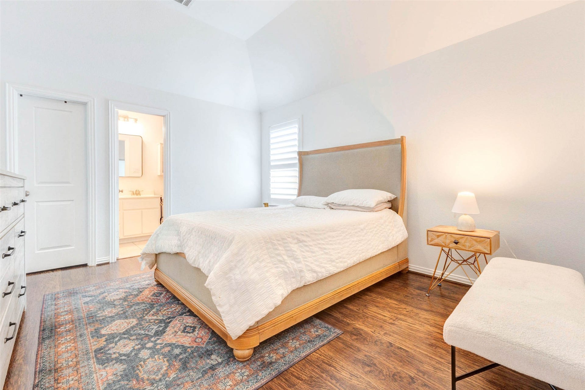 This spacious bedroom offers a serene and airy feel with its high ceilings and soft neutral finishes. Rich wood flooring adds warmth, while the open layout provides flexibility for furniture placement. Natural light streams in, enhancing the peaceful ambiance. With convenient access to a nearby bathroom, this room offers comfort and convenience in a well-designed space