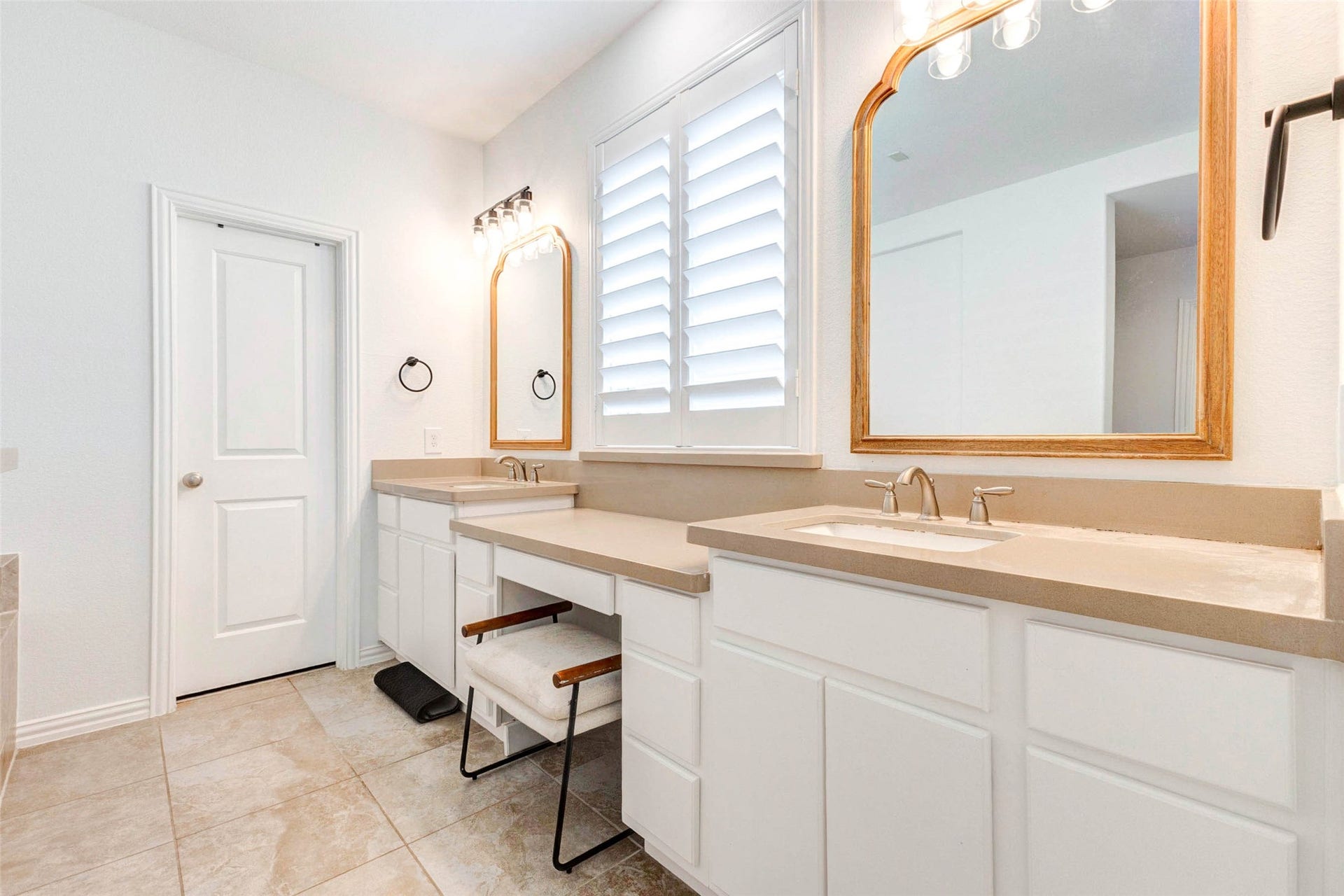 This spacious primary bathroom features dual vanities with abundant counter space, offering both style and functionality. Clean cabinetry, updated fixtures, and large framed mirrors create a bright and polished look. A dedicated sitting area adds convenience, while the window with plantation shutters brings in soft natural light. Designed for comfort and efficiency, this space makes daily routines feel elevated and organized.