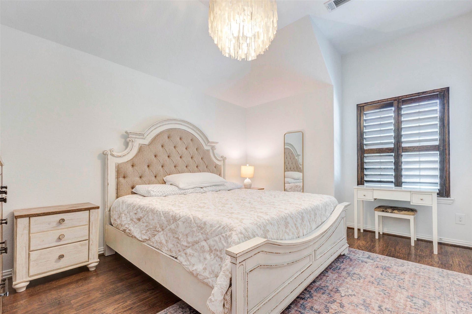 This inviting secondary bedroom blends comfort and charm with its elegant upholstered headboard and soft neutral palette. Warm wood flooring grounds the space, while natural light filters through plantation shutters, creating a bright yet cozy atmosphere. A chandelier adds a touch of sophistication, and the room offers space for both function and relaxation.