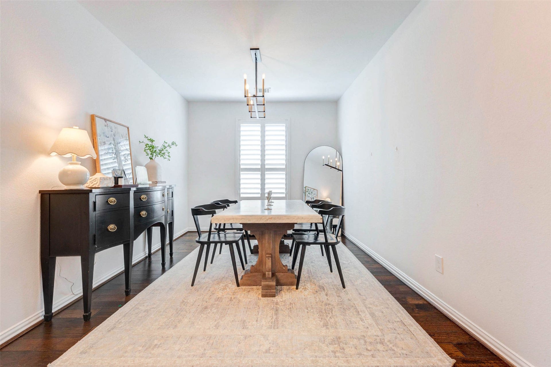 This elegant formal dining room sets the stage for memorable gatherings, featuring a modern light fixture and a spacious layout for entertaining. Rich wood flooring adds warmth, while the large window with plantation shutters allows natural light to fill the space. A stylish sideboard and clean lines create a sophisticated yet welcoming environment, perfect for both intimate dinners and larger celebrations.