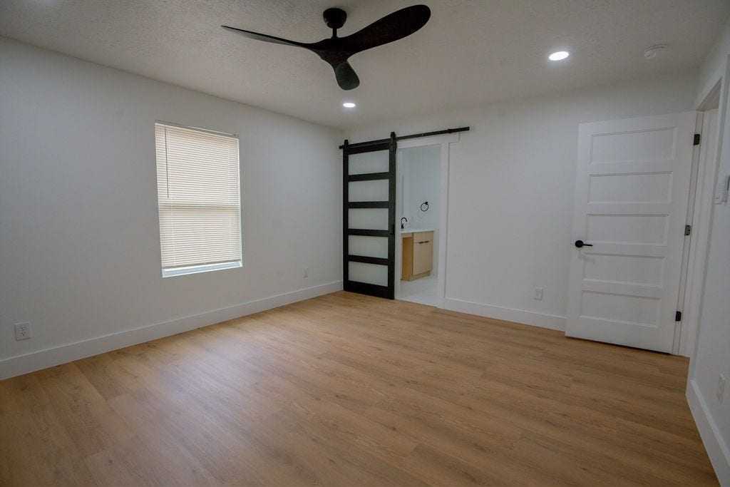 Spacious bedroom featuring sleek wood style flooring, a modern black ceiling fan, and a stylish sliding barn door leading to an en-suite bathroom.