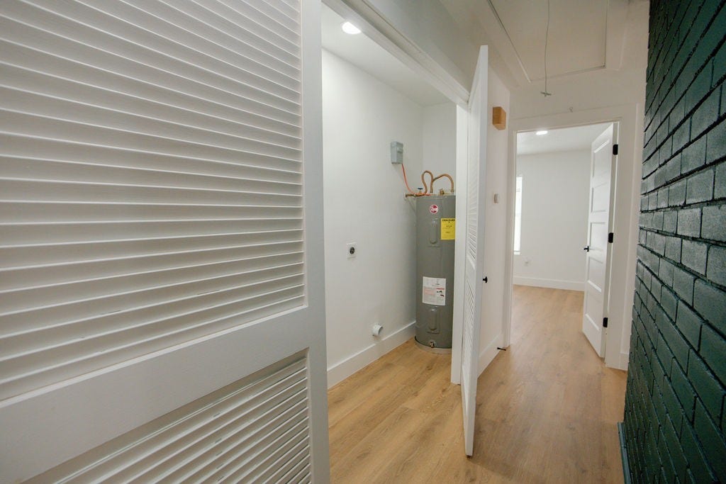 A utility closet with a water heater is tucked into a bright hallway featuring sleek wood style flooring and a stylish dark brick accent wall.