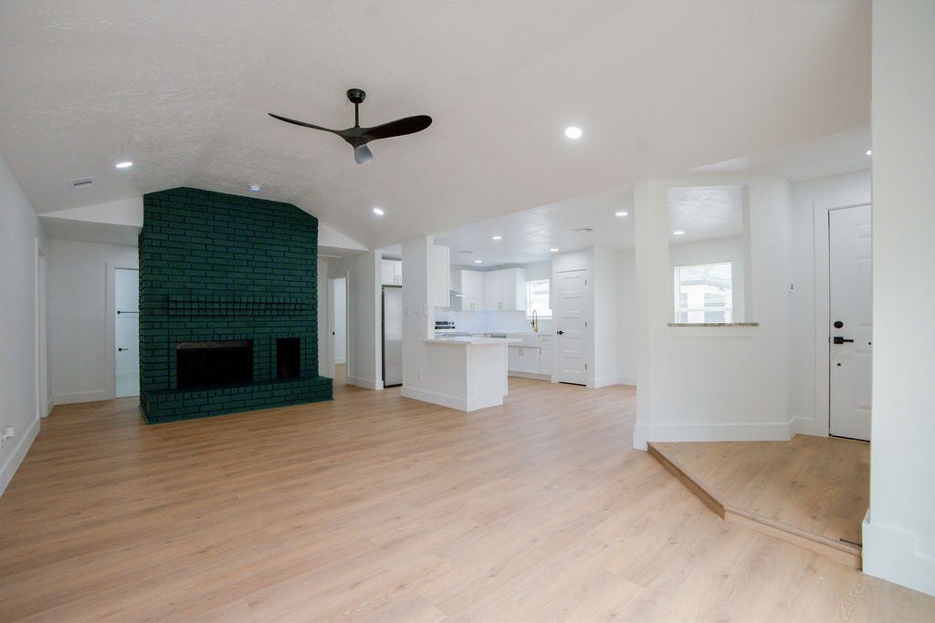 Discover a spacious open-plan living area featuring a striking dark green brick fireplace, modern ceiling fan, and seamless flow into a bright, updated kitchen with sleek finishes.