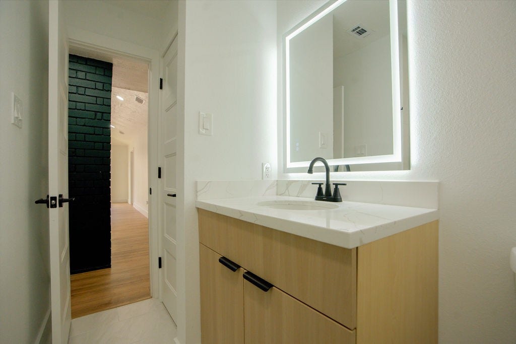 Modern bathroom with a sleek vanity, illuminated mirror, and elegant fixtures, leading to a bright hallway with wood style flooring.