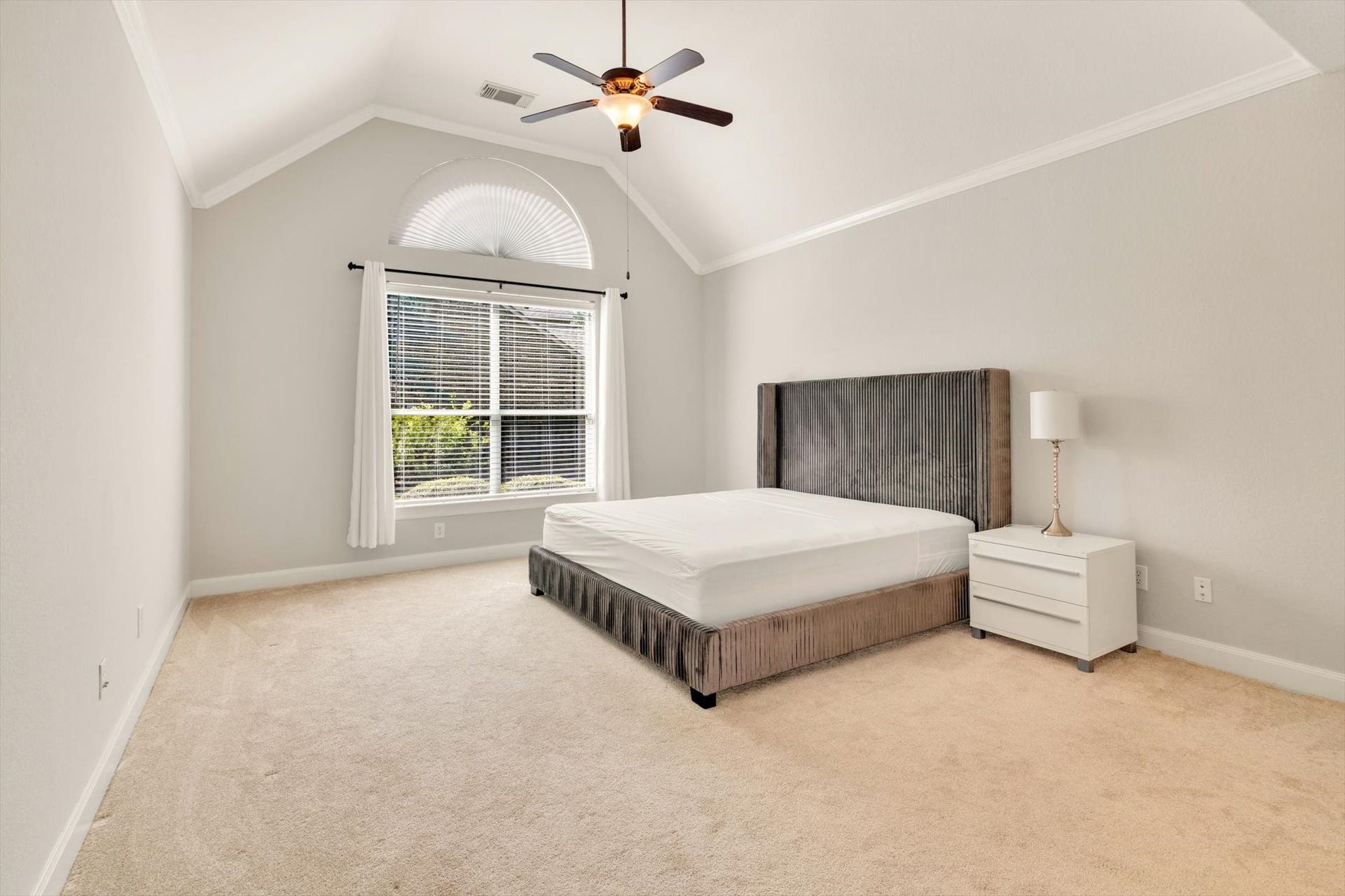 Primary bedroom features a high vaulted ceiling and neutral carpeting. It includes a large window with elegant drapes, allowing ample natural light. The room is furnished with a stylish bed and nightstand, creating a cozy and inviting atmosphere.