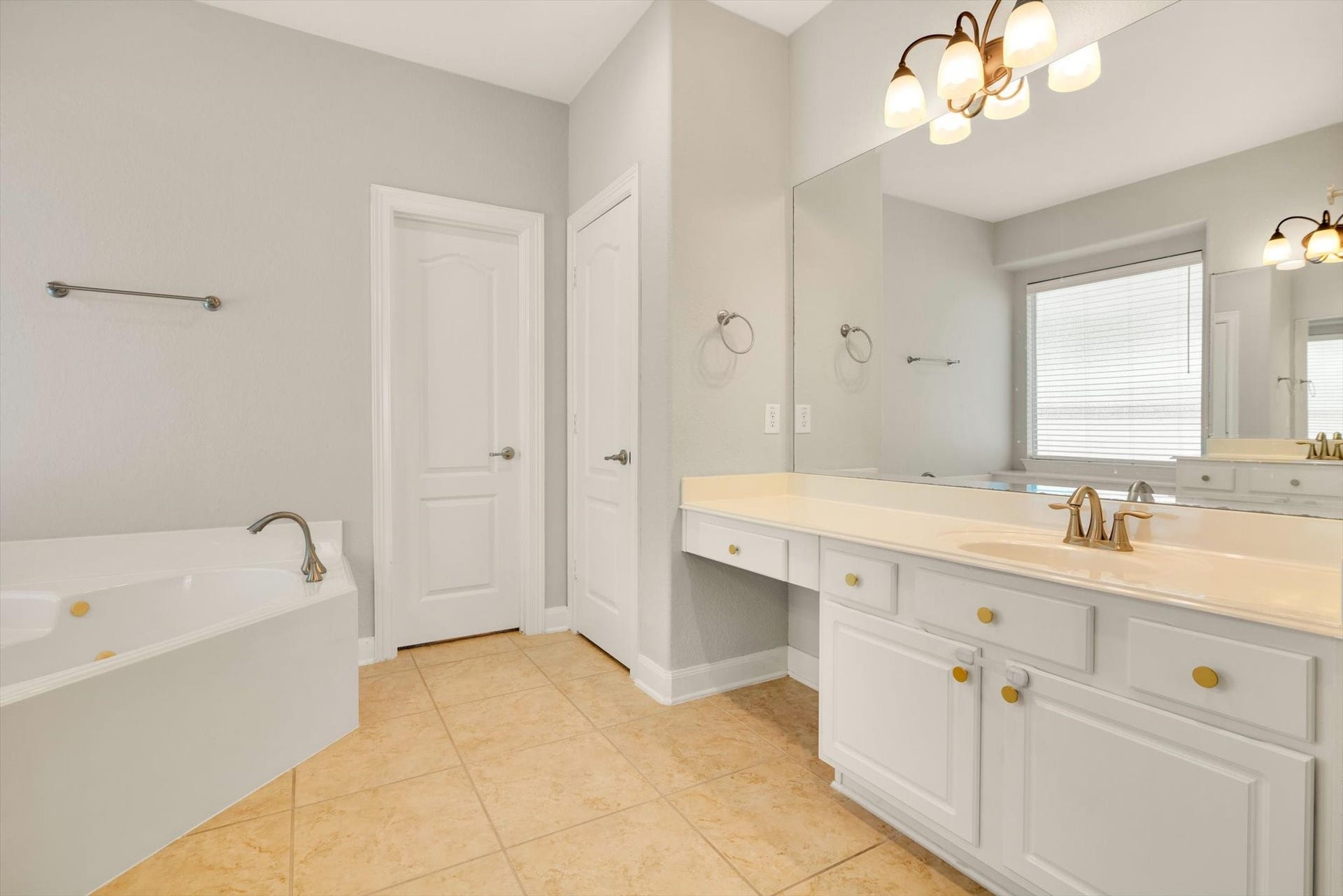 Primary bathroom features a spacious layout with a large mirror, dual sinks, elegant lighting fixtures, a soaking tub, and neutral tile flooring, creating a bright and inviting space.