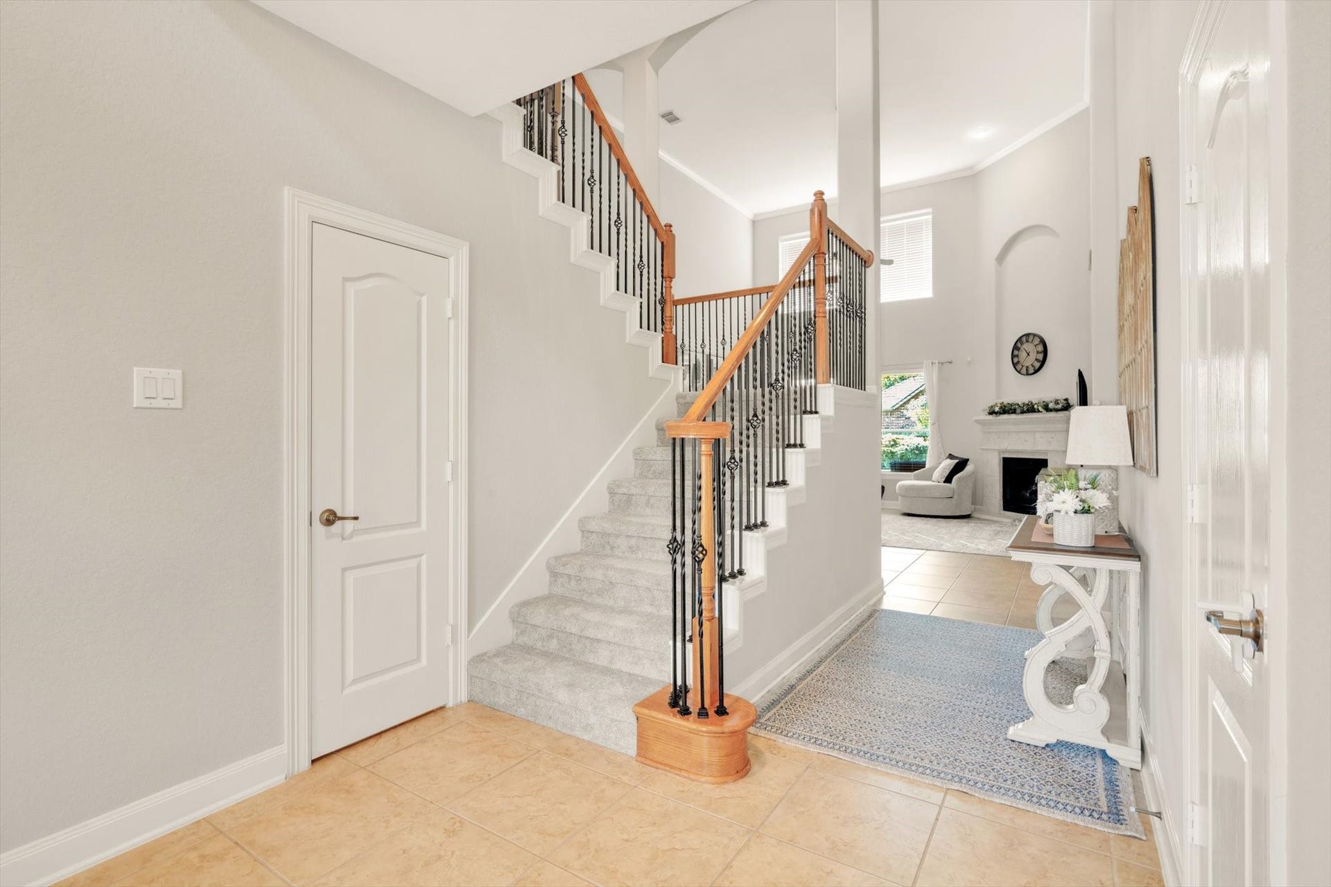 Bright entryway features a tiled floor and a carpeted staircase with wooden handrails and wrought iron spindles. The open layout leads into a high-ceilinged living area with a fireplace and large windows, creating a welcoming and airy atmosphere.