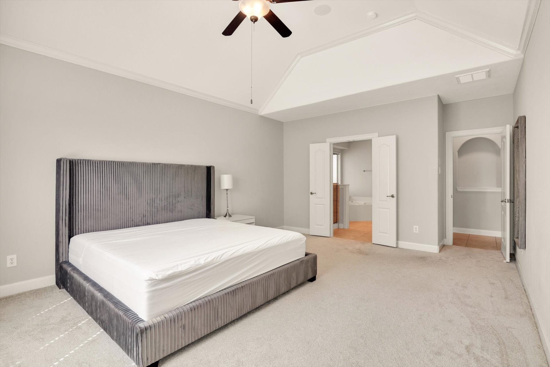 Primary bedroom features neutral tones and plush carpeting, with a stylish upholstered bed. It includes a ceiling fan and convenient ensuite access through double doors, leading to a bathroom with a soaking tub. Ideal for comfort and relaxation.