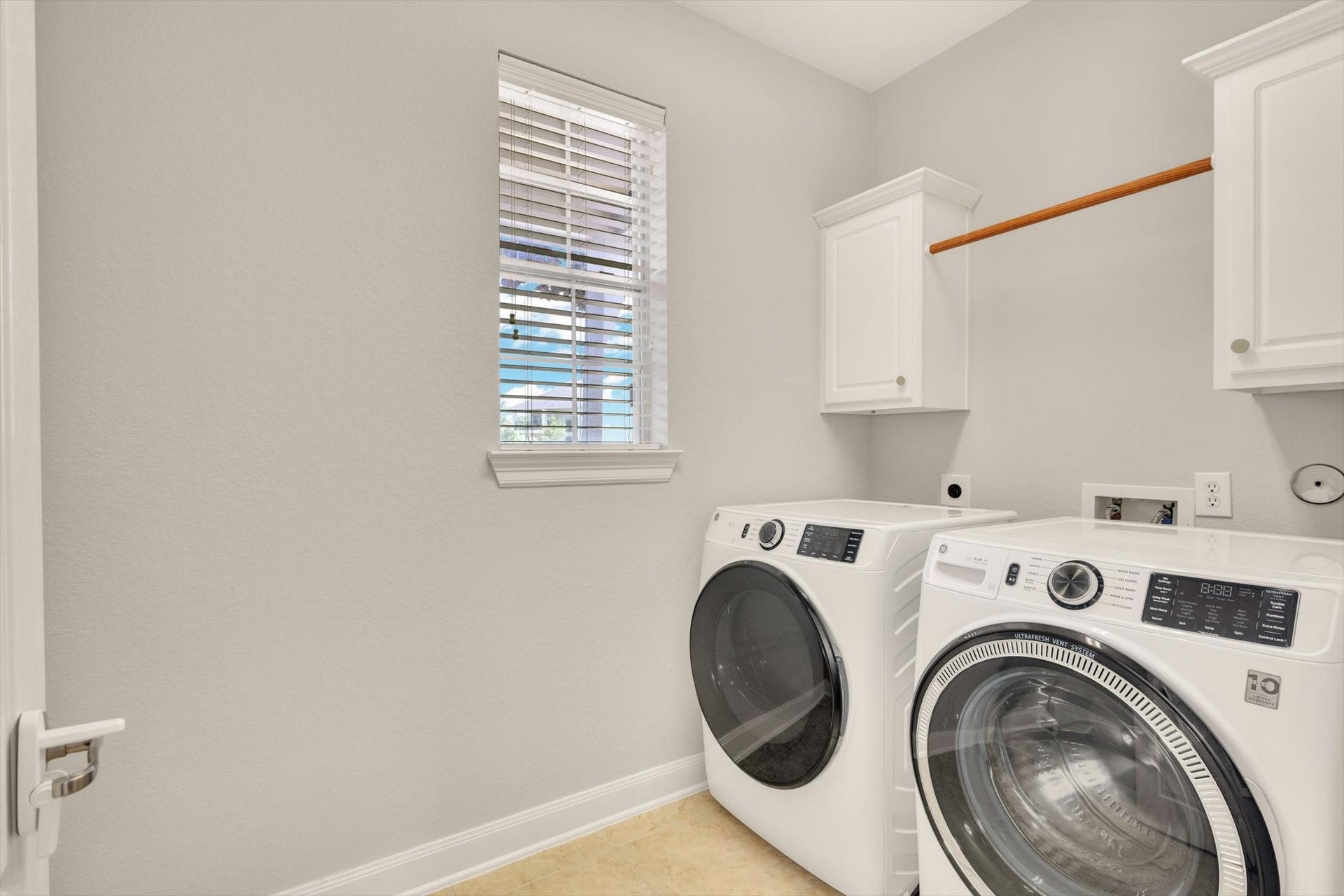 This is a bright, modern laundry room featuring a washer and dryer set, white cabinetry for storage, a hanging rod, and a window for natural light.