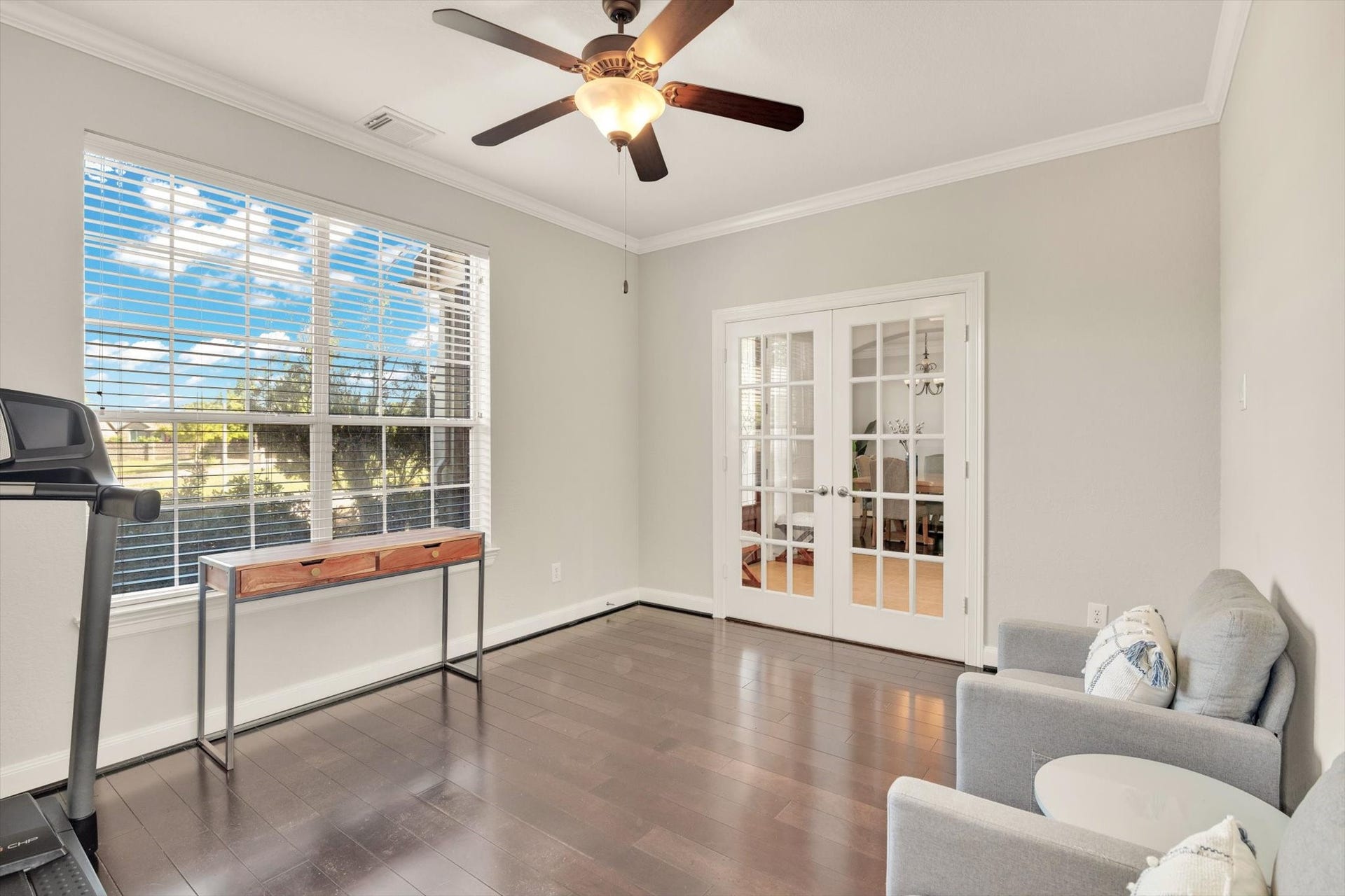 This bright room features dark hardwood floors and a ceiling fan, with large windows offering plenty of natural light. French doors open to an adjoining space, creating an inviting and versatile area for an office or sitting room.