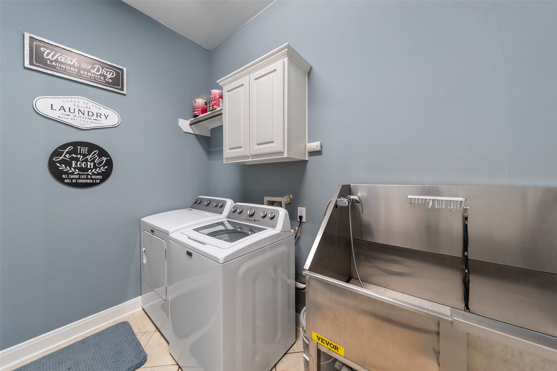 34 Danby Place, The Woodlands, TX 77375 This spacious laundry room features a washer and dryer, ample cabinetry for storage, and a large utility sink. The walls are decorated with charming laundry-themed signs, creating a welcoming and functional atmosphere.