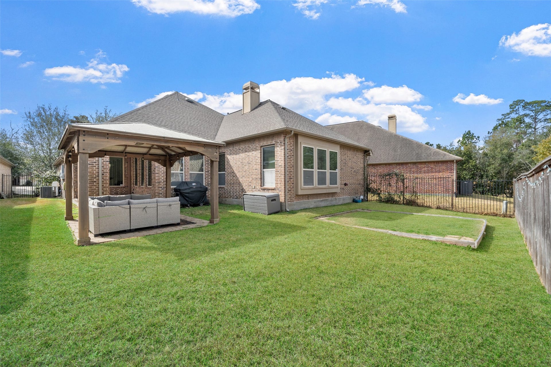 34 Danby Place, The Woodlands, TX 77375 Spacious backyard with a well-maintained lawn, featuring a covered seating area perfect for outdoor relaxation and entertainment. The brick exterior adds charm and durability. The yard is spacious, with green grass and a small paved area, providing a cozy and inviting outdoor setting.