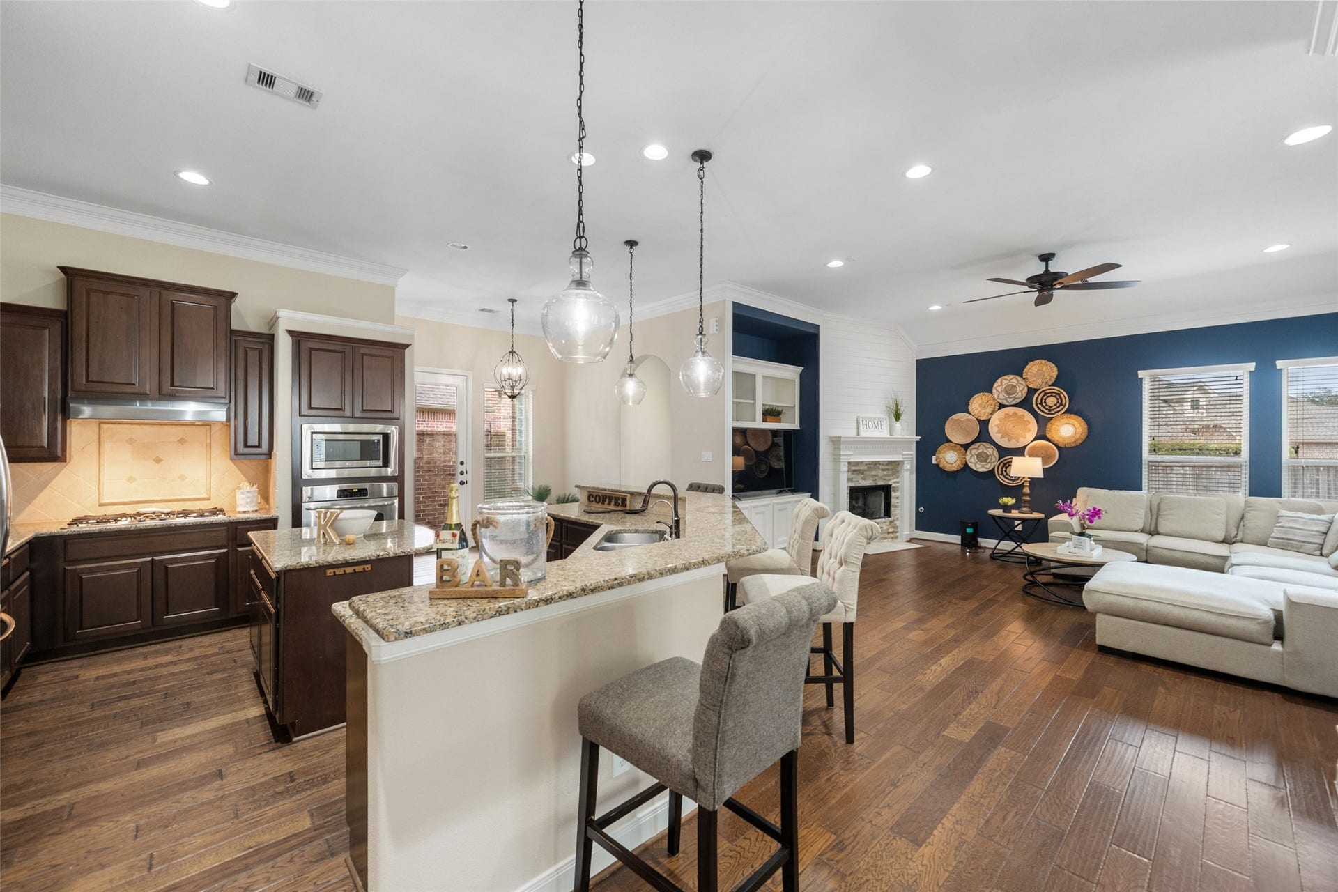 34 Danby Place, The Woodlands, TX 77375 A modern open-concept living area featuring a kitchen, living room with a cozy fireplace, and a stylish accent wall with decorative art. A large sectional sofa offers ample seating, creating an inviting and functional space, ideal for relaxation and entertaining.
