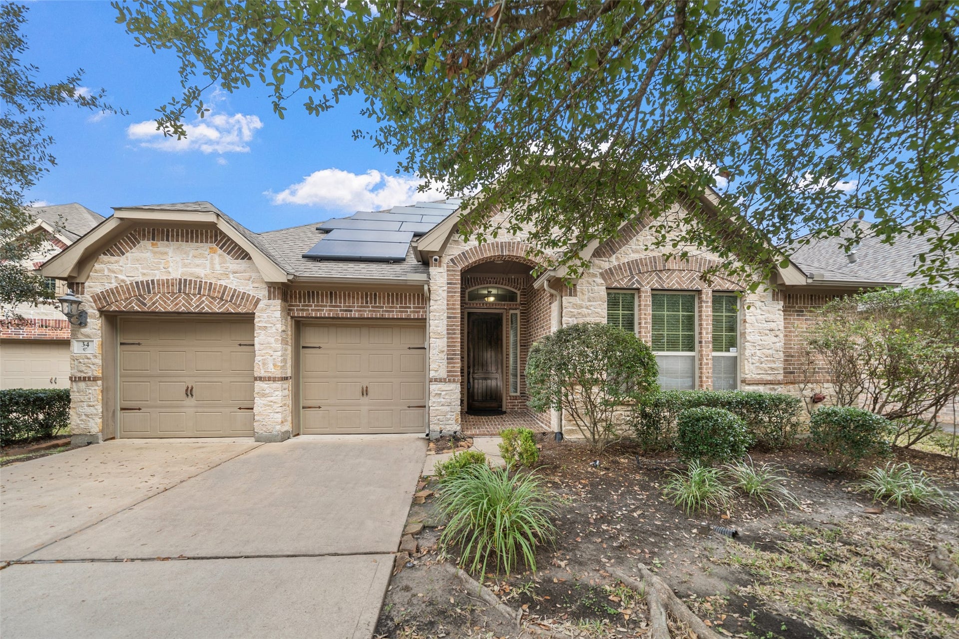 34 Danby Place, The Woodlands, TX 77375 This charming single-story home features a brick and stone exterior, a two-car garage, and a welcoming entryway. The front yard is landscaped with shrubs and trees, and solar panels are installed on the roof, enhancing energy efficiency.
