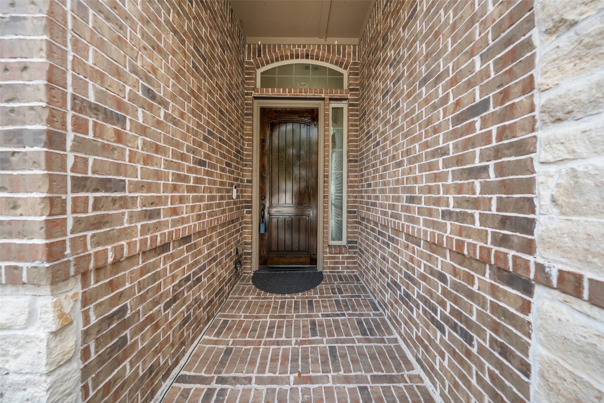 34 Danby Place, The Woodlands, TX 77375 A welcoming entryway with a rich wooden front door, surrounded by arched brick walls. The brickwork extends to the floor, creating a cohesive and inviting entrance.