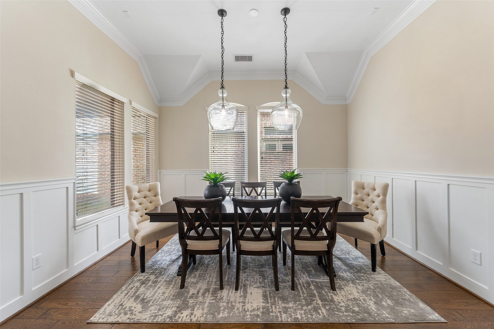 34 Danby Place, The Woodlands, TX 77375 This elegant dining room features a vaulted ceiling, soft neutral walls, and white wainscoting. It has a large window for natural light, a stylish dark wood table with cushioned chairs, and modern pendant lighting.